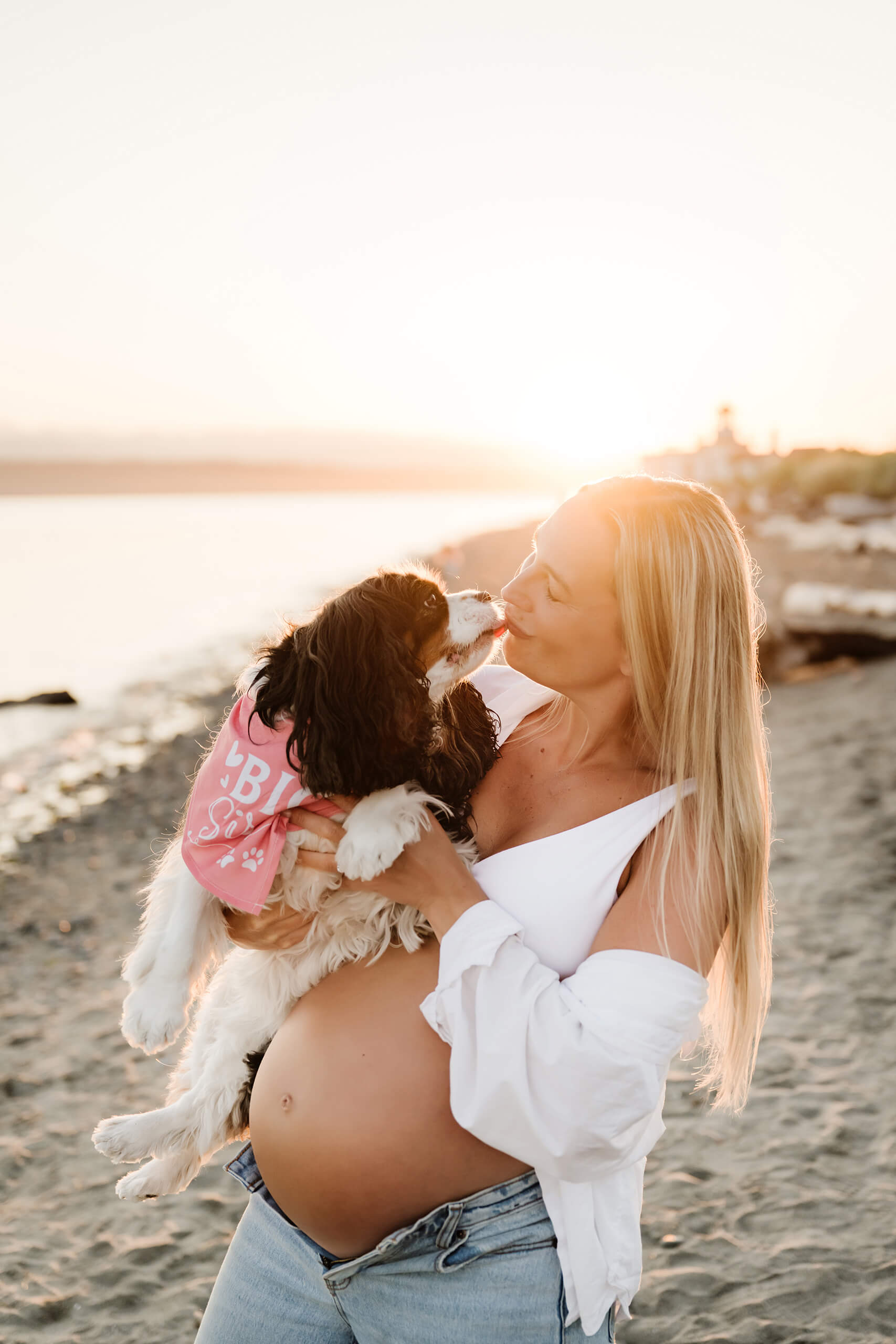 Pregnant woman smiling as her dog kisses her during a golden-hour maternity photoshoot by the water.