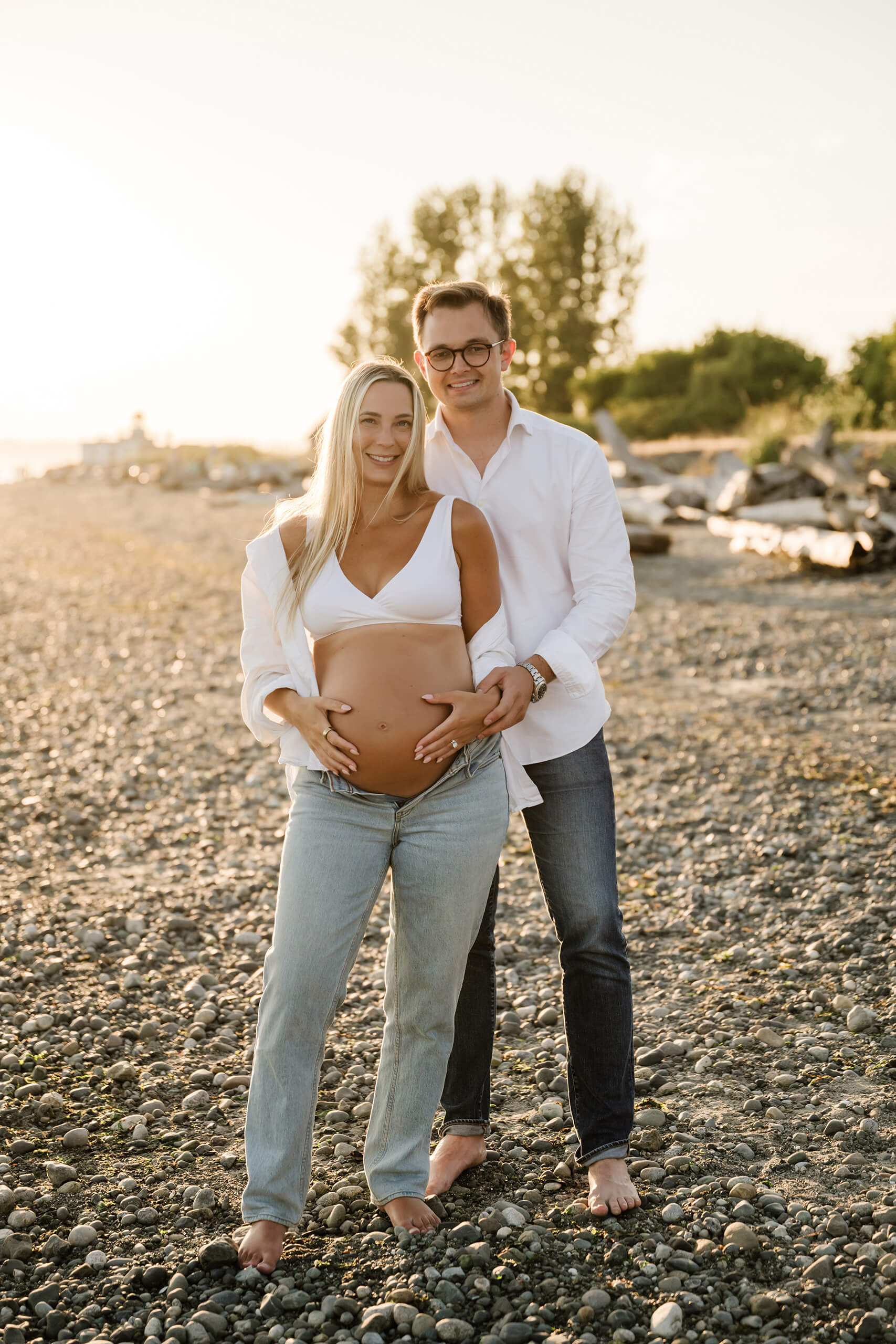 Pregnant couple standing barefoot on a rocky beach during a sunset maternity session near Seattle.