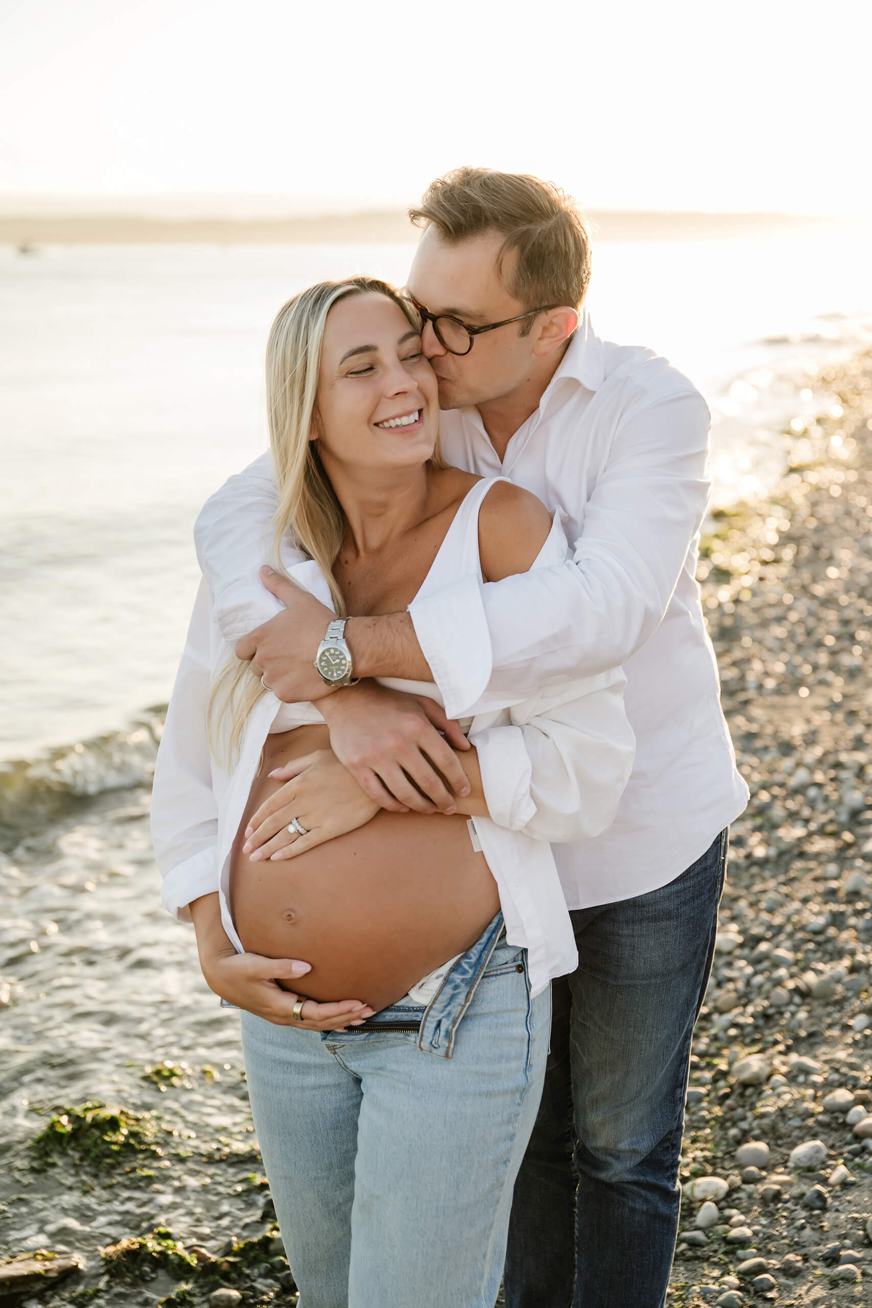 Expecting father embracing and kissing his pregnant partner on a beach at golden hour.