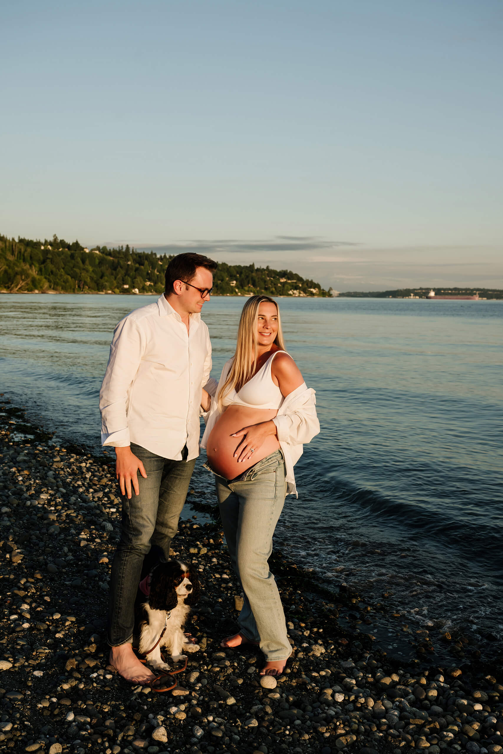 Expecting couple with their dog standing along the shoreline during a waterfront maternity session.