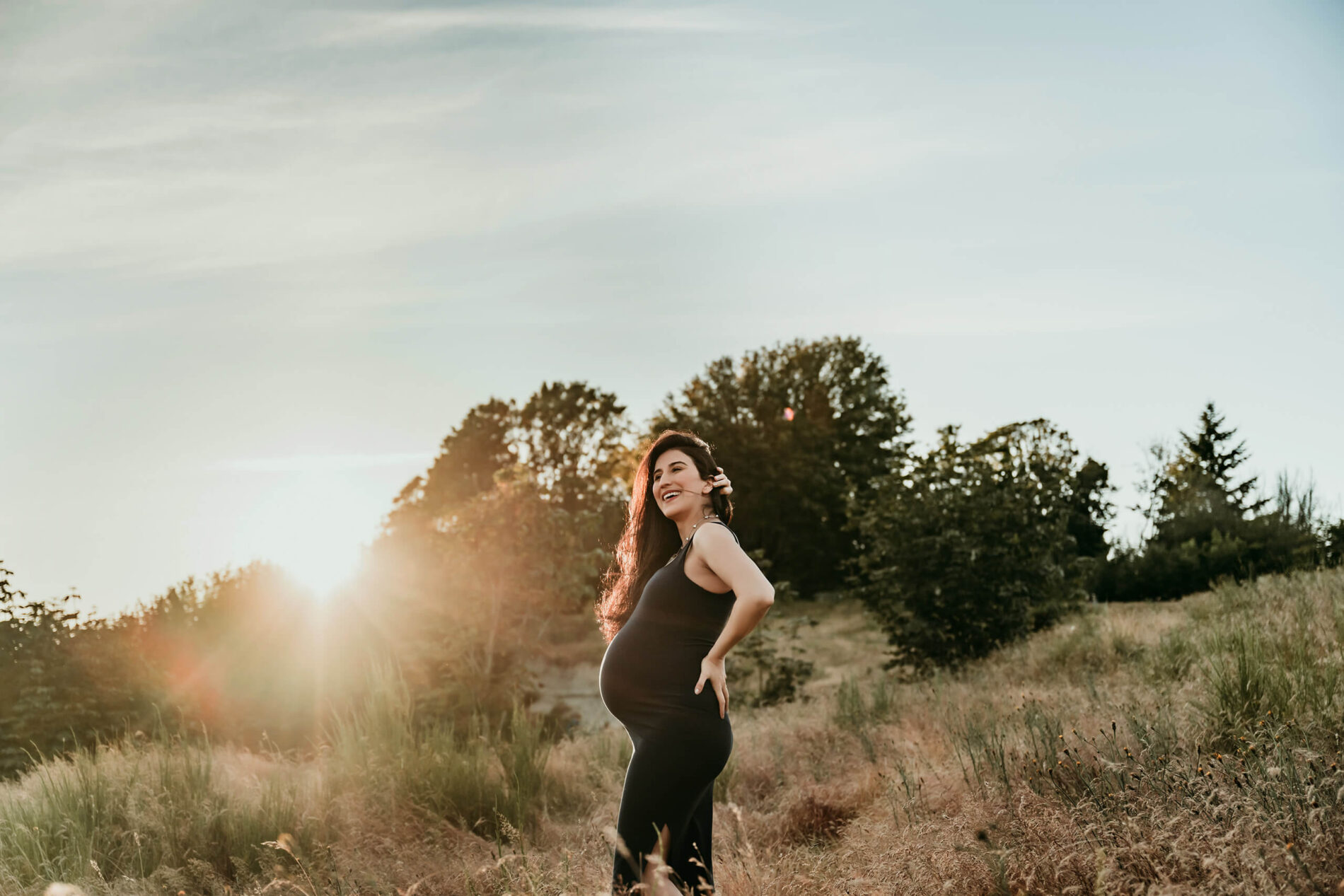 Pregnant woman posing in a black dress in a golden field during a sunset maternity session.