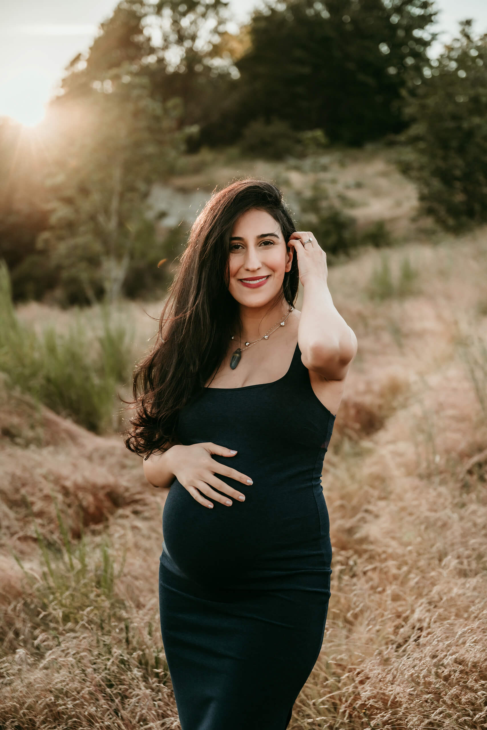 Pregnant woman in a dark dress standing in tall grass during an outdoor maternity portrait.