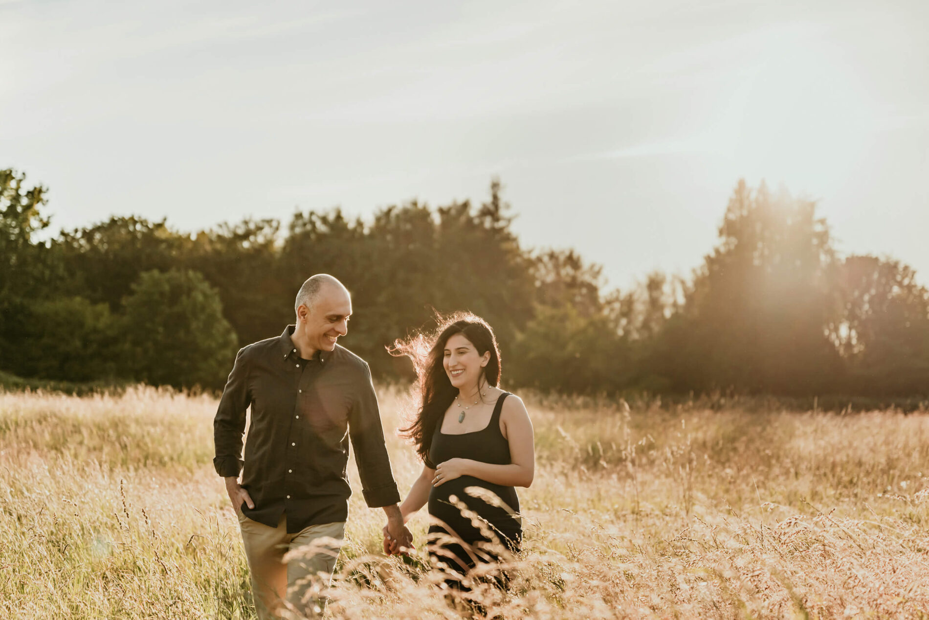 Expecting couple walking hand in hand through a golden field during a sunset maternity session.