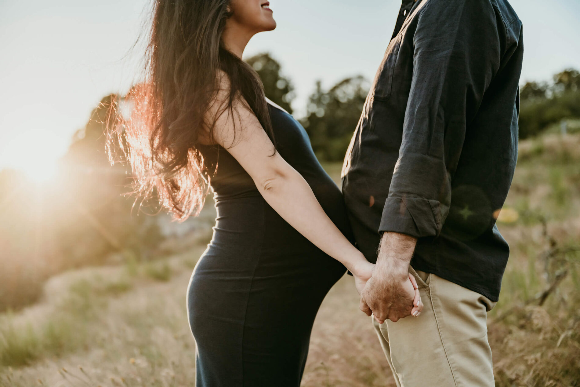 Close-up of a pregnant couple holding hands in a grassy field at golden hour.