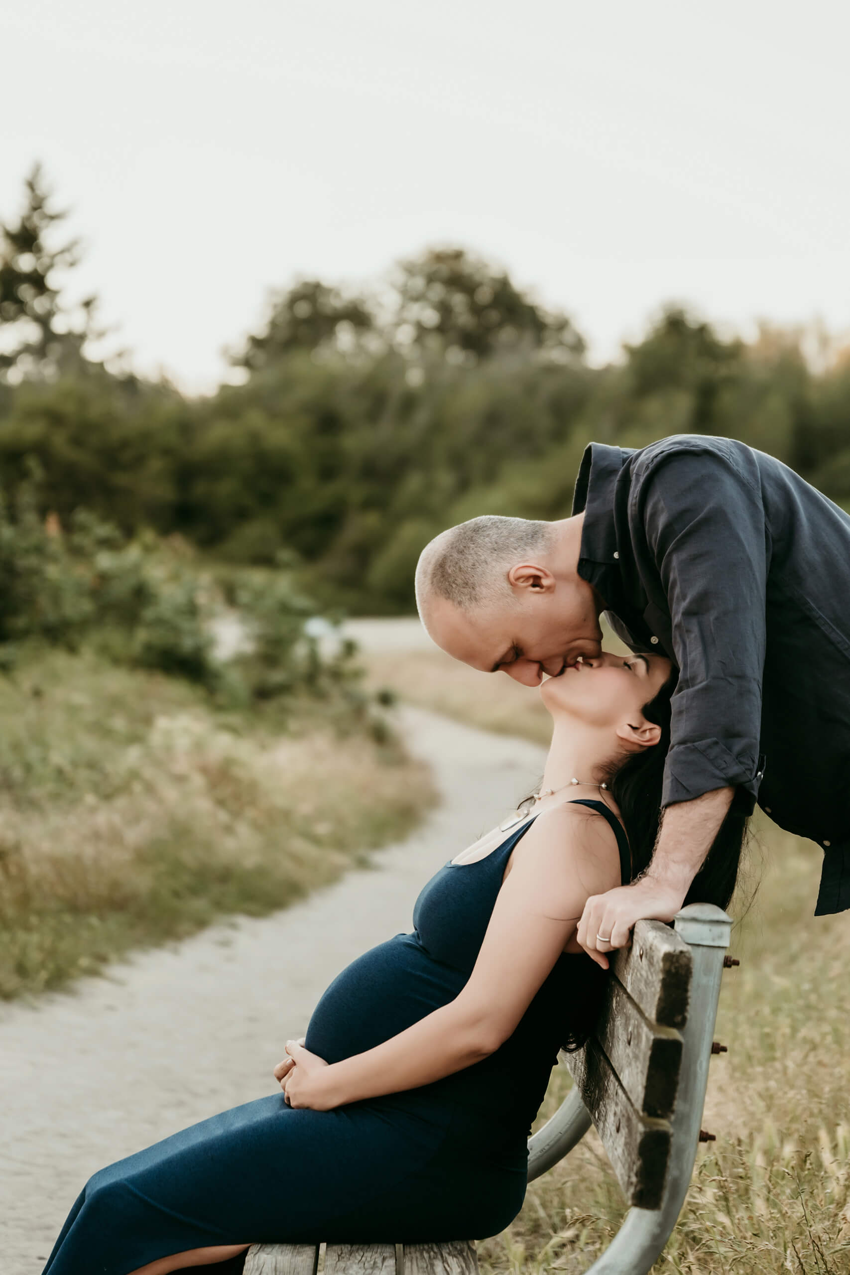 Pregnant woman seated on a wooden bench while her partner leans in for a kiss.