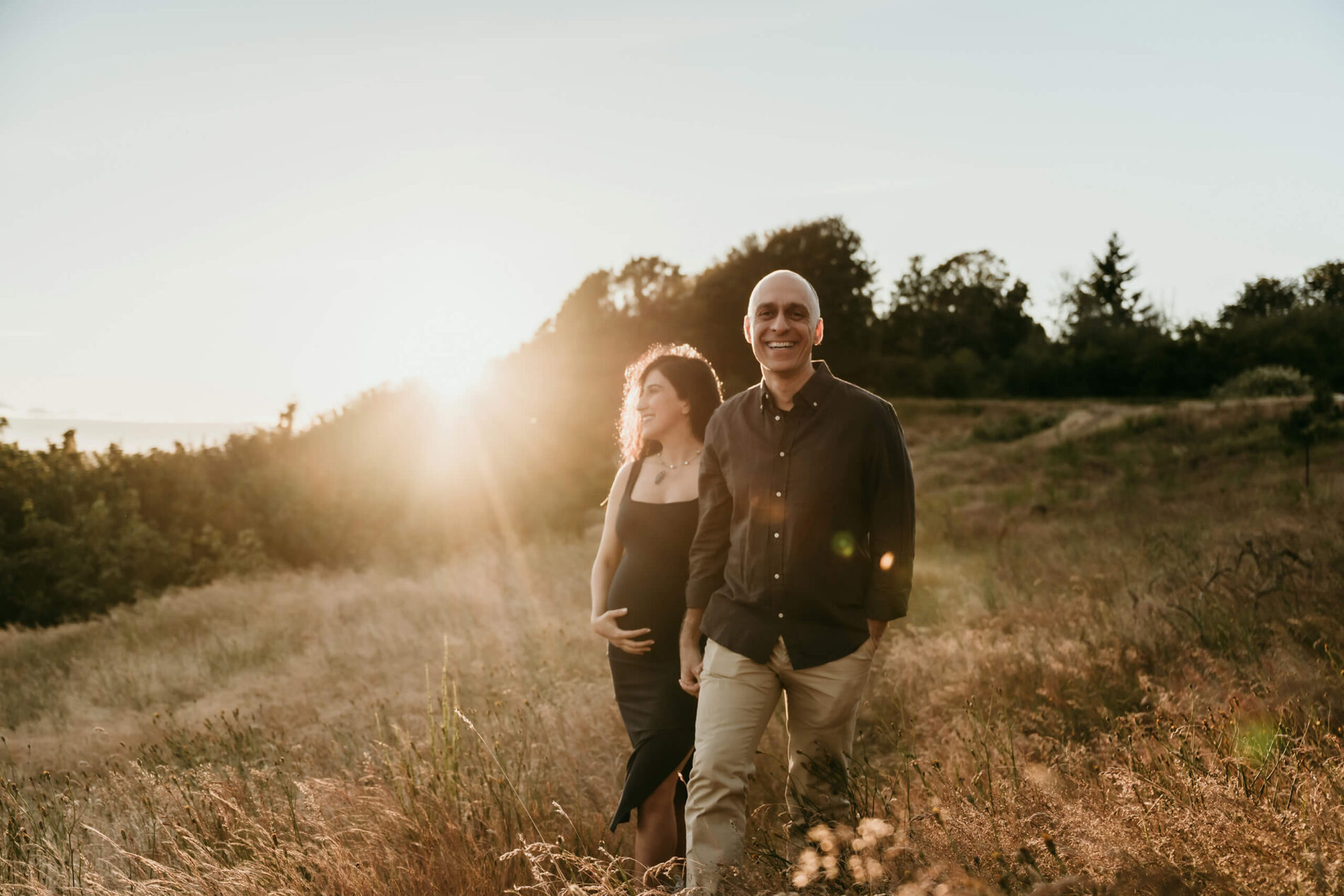 Expecting couple walking through a sunlit field during an outdoor maternity session.