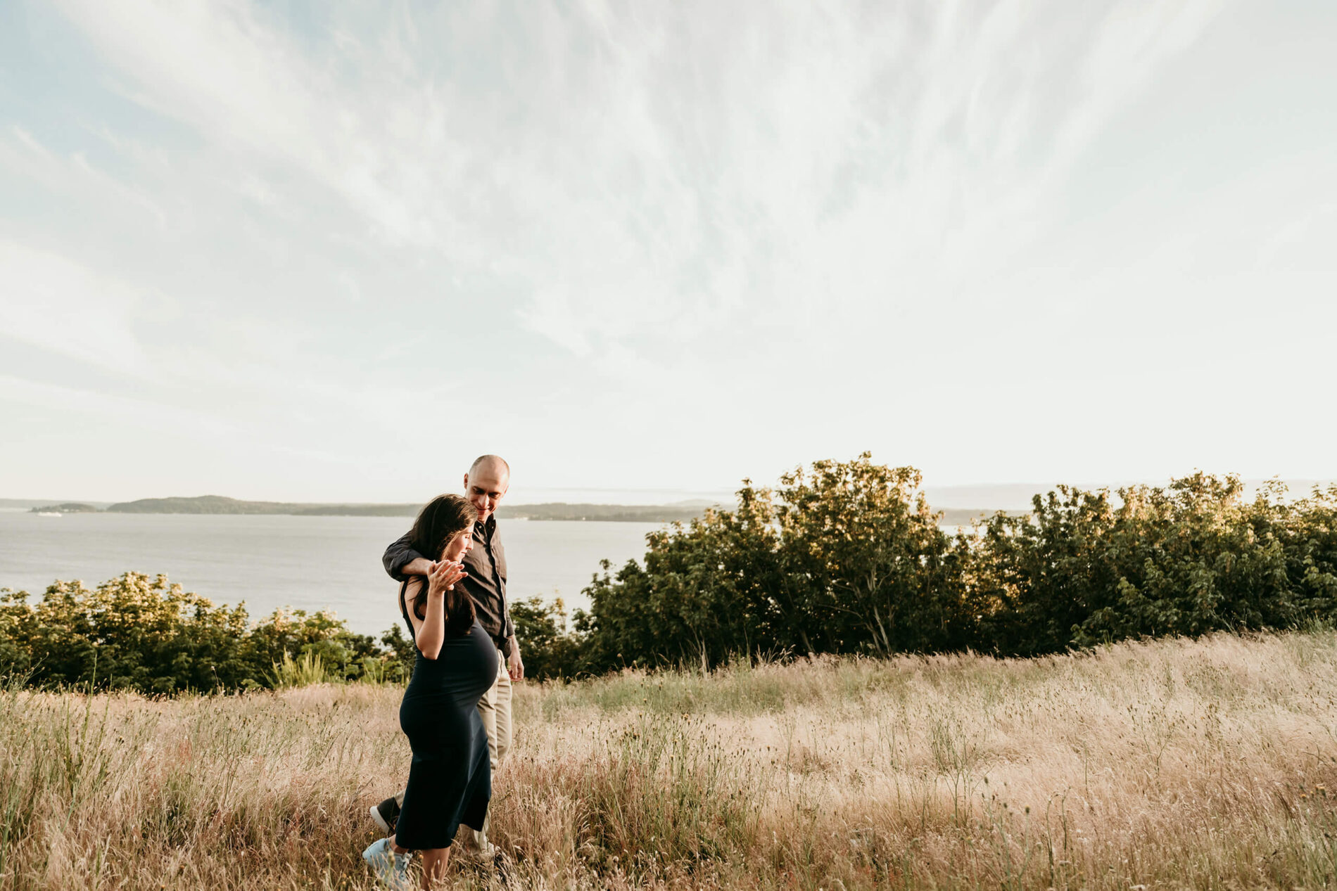 Pregnant couple walking together in a grassy field overlooking the water.