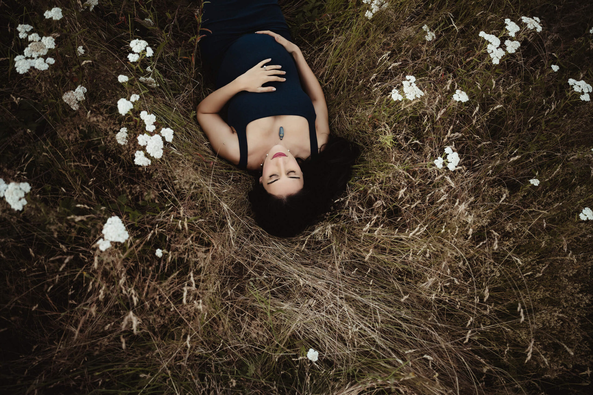 Pregnant woman lying in tall grass and white flowers during an outdoor maternity portrait.