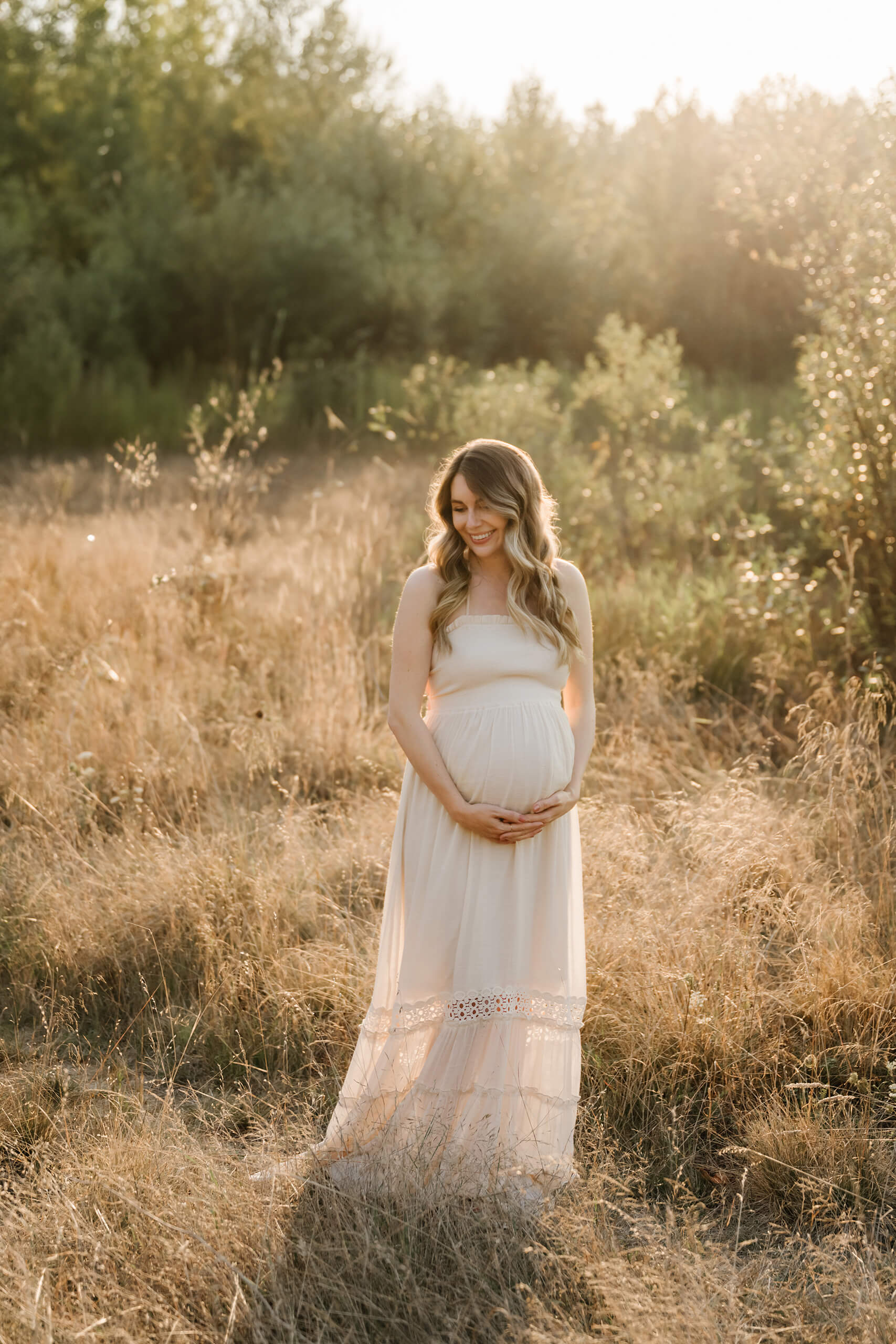 Pregnant woman in a white dress standing in a golden field during an outdoor maternity session.