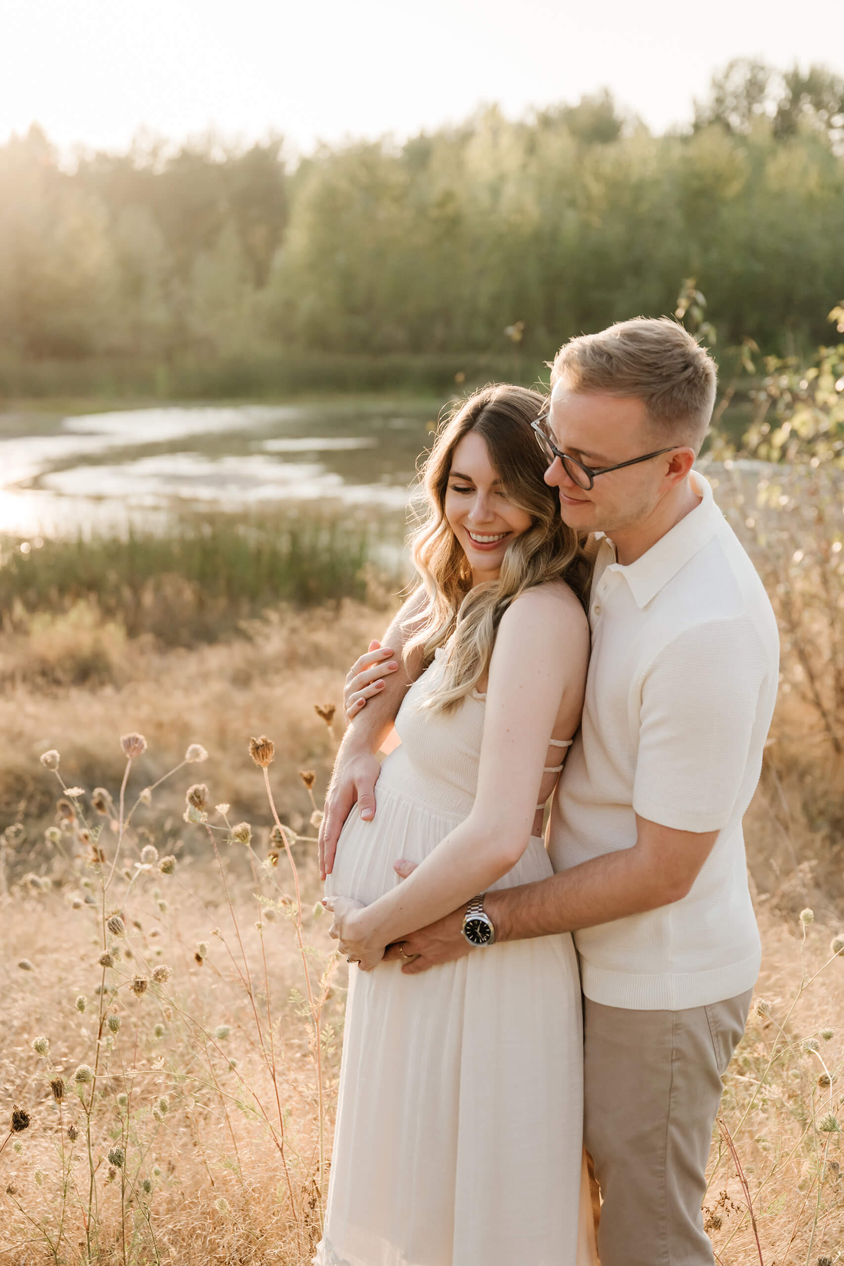 Expecting couple embracing in a golden field near the water during an outdoor maternity photoshoot.