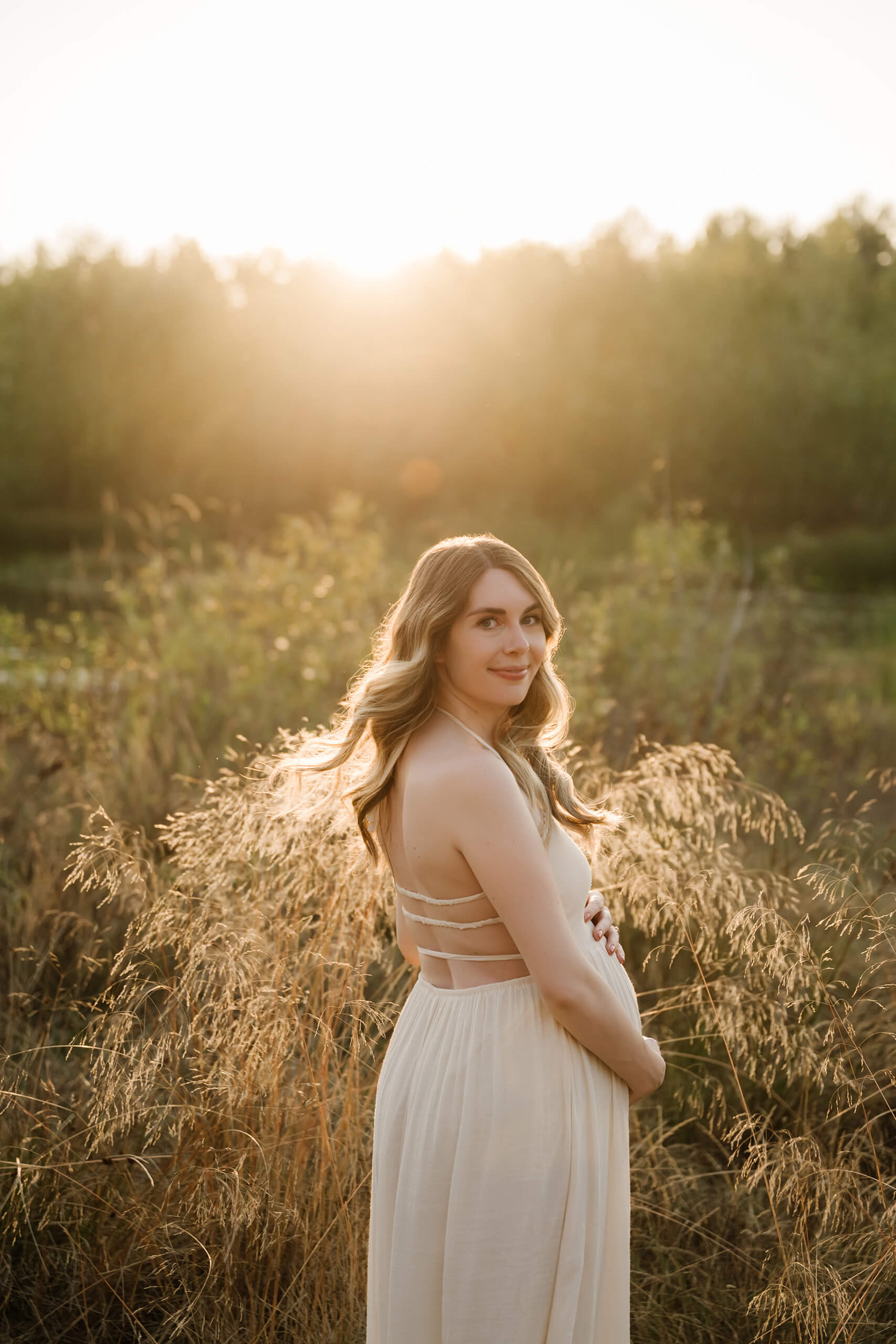 Pregnant woman in a white dress looking back in a golden field at sunset.