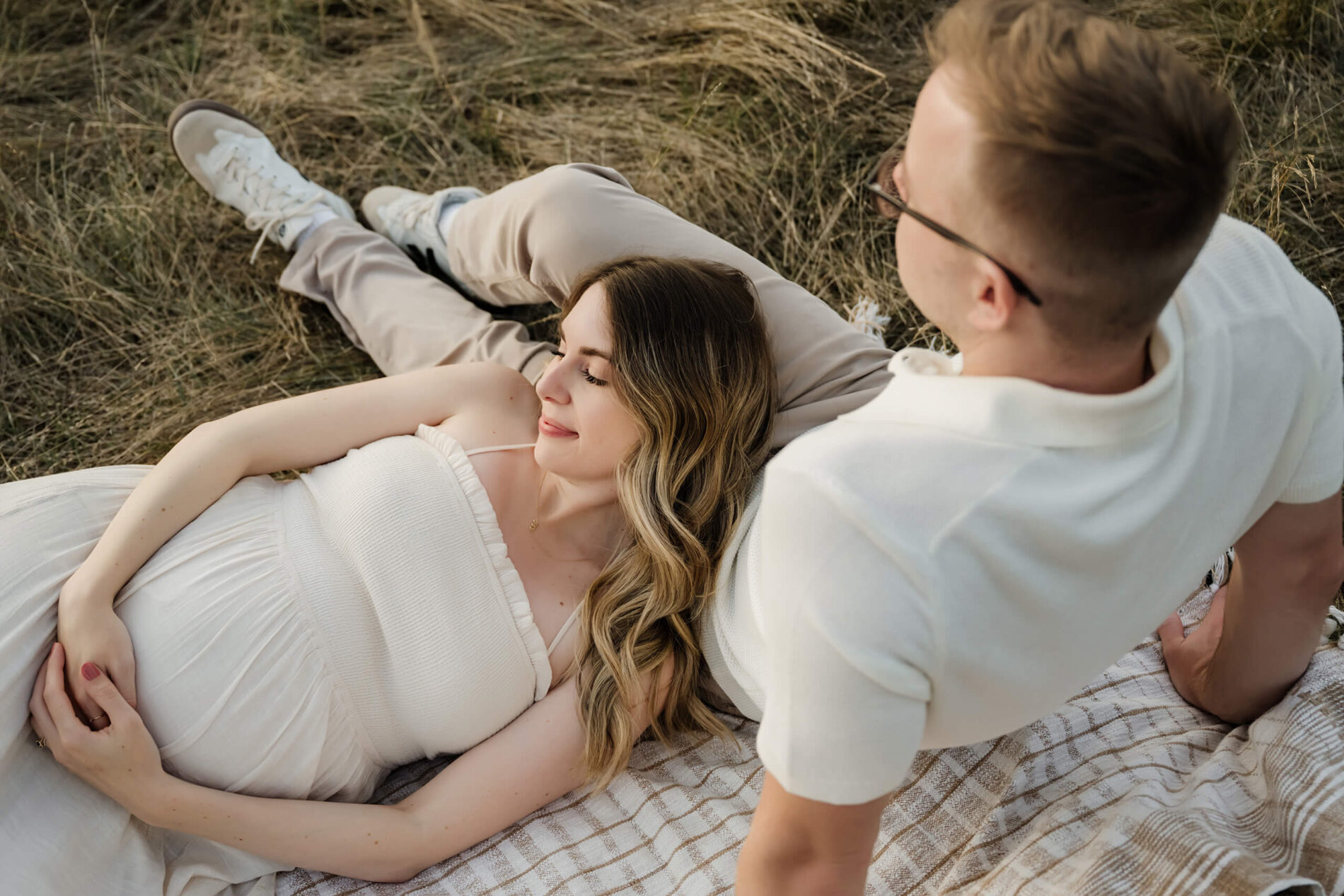 Pregnant woman resting on a blanket in a field during a soft outdoor maternity portrait.