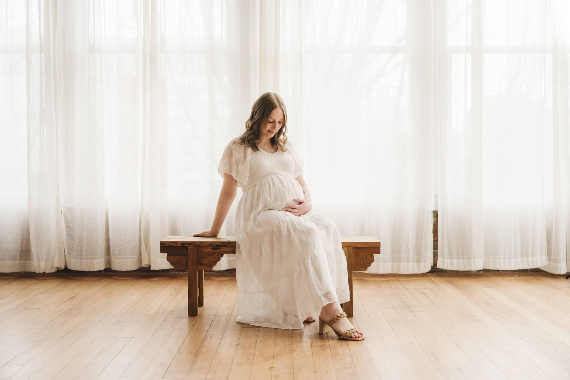 Pregnant woman in a white dress seated on a wooden bench in a bright studio with sheer curtains.