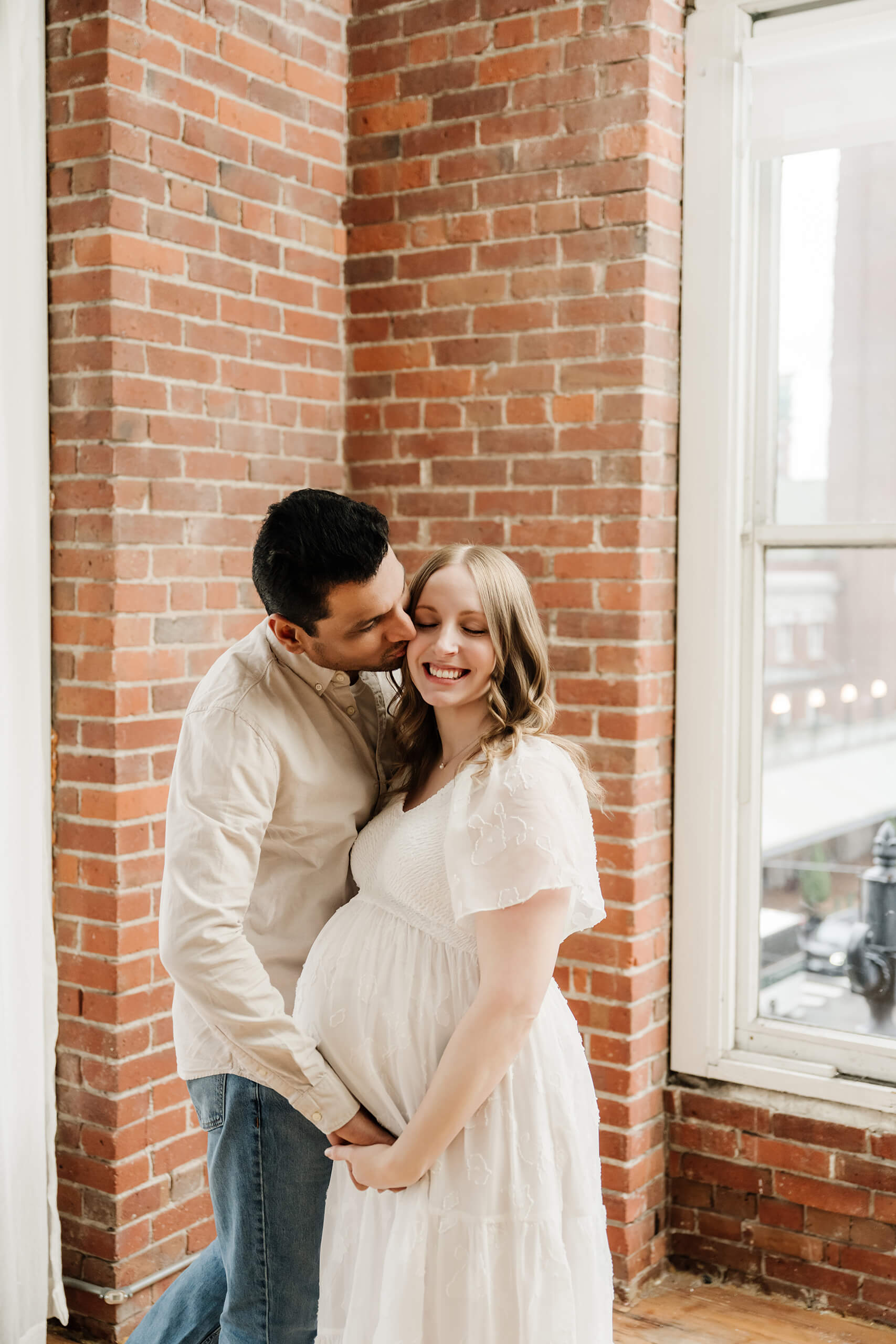 Expecting couple standing together by a brick wall and window during a studio maternity session.