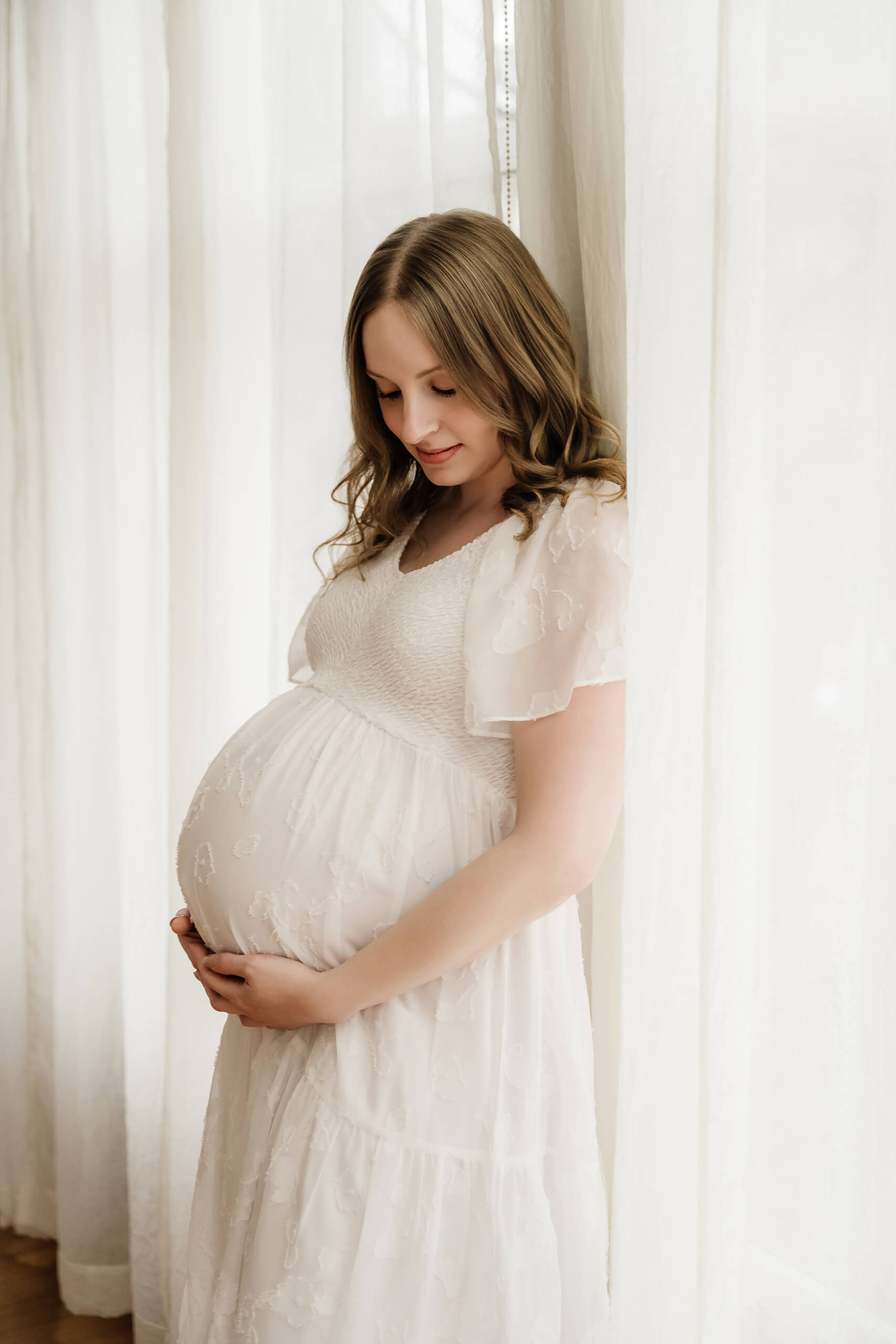 Pregnant woman in a white dress standing near sheer curtains with her hands on her belly.