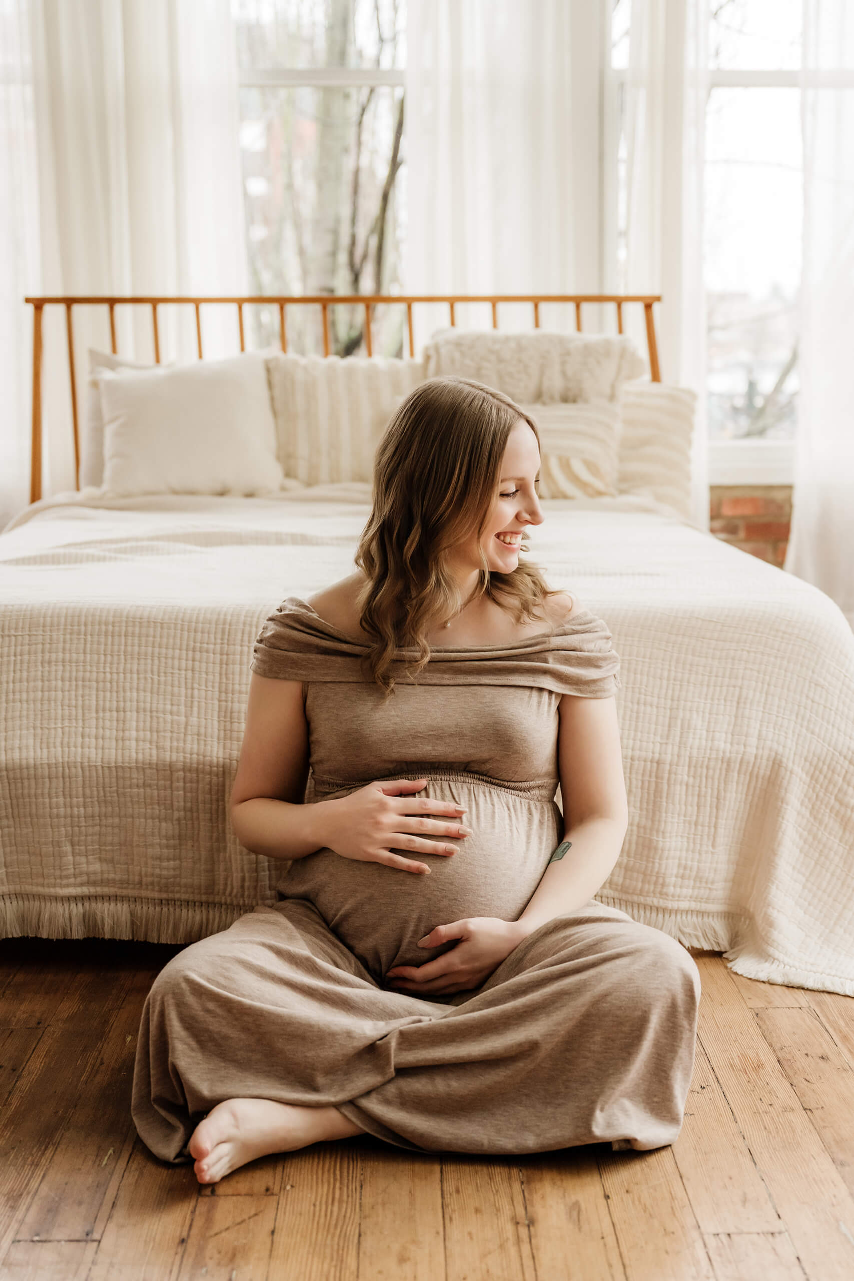 Pregnant woman seated on the floor in front of a bed for a soft studio maternity portrait.