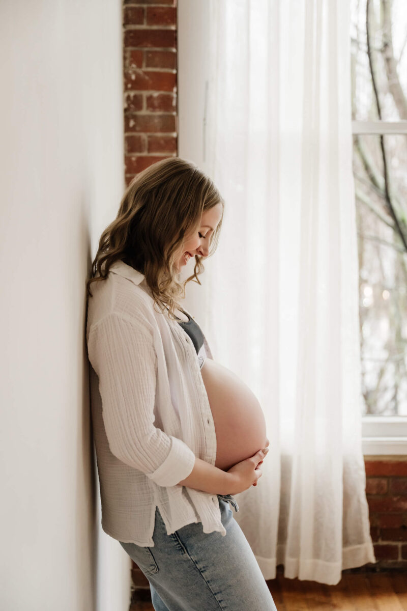 Pregnant woman standing by a window with her hands on her belly in a bright studio setting.
