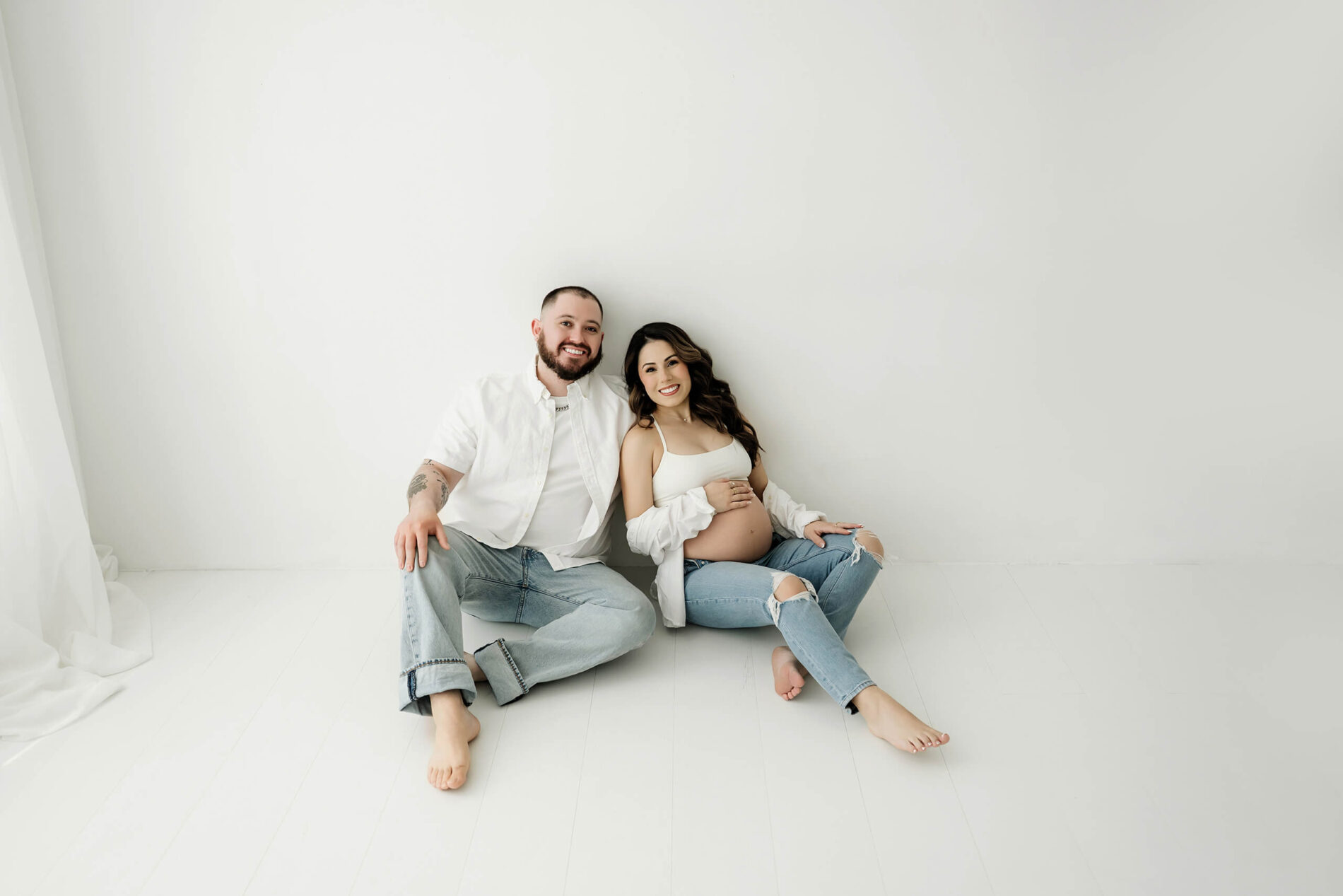Expecting couple seated together on the floor against a simple white studio background.