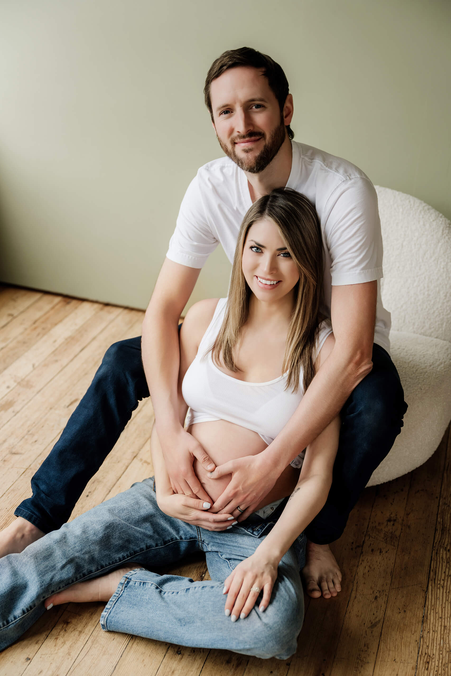 Expecting couple seated together on a wood floor with their hands resting on the baby bump.
