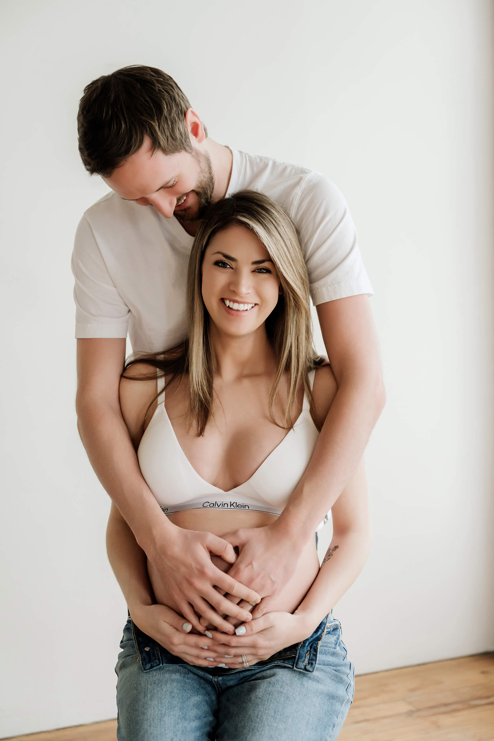Expecting couple smiling together in a bright studio with their hands on the baby bump.