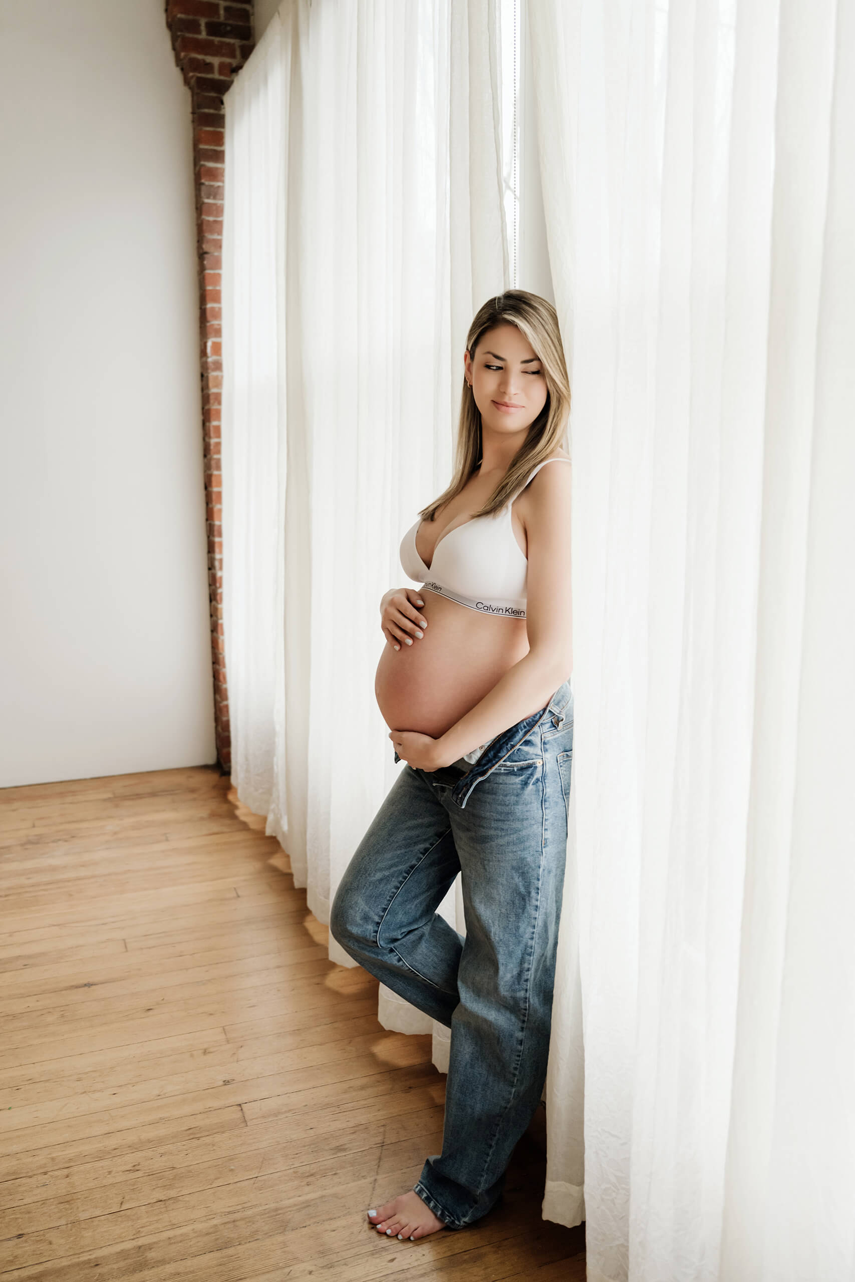 Pregnant woman standing beside sheer curtains for a simple studio maternity portrait.