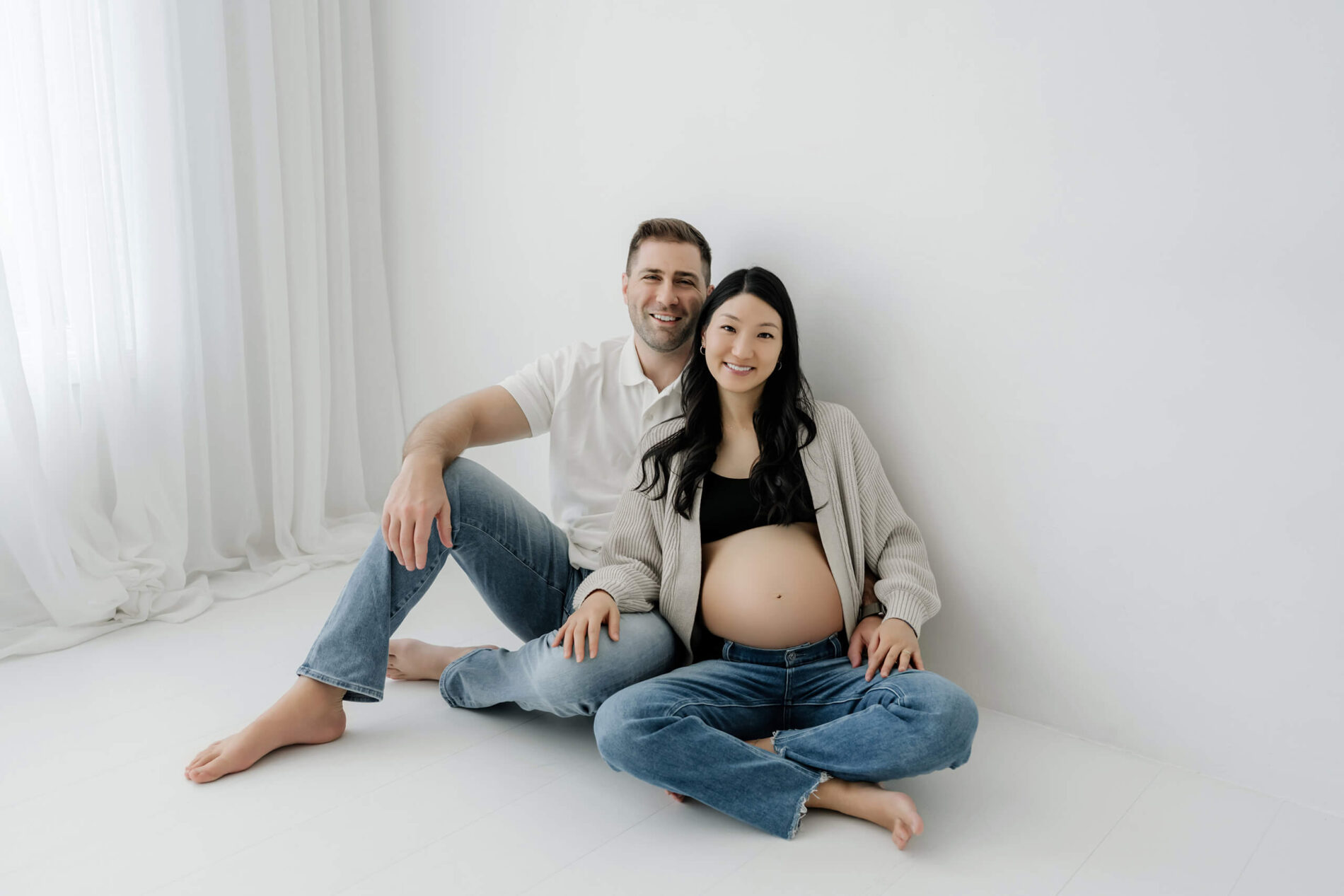 Expecting couple seated together in a bright white studio during a relaxed maternity session.
