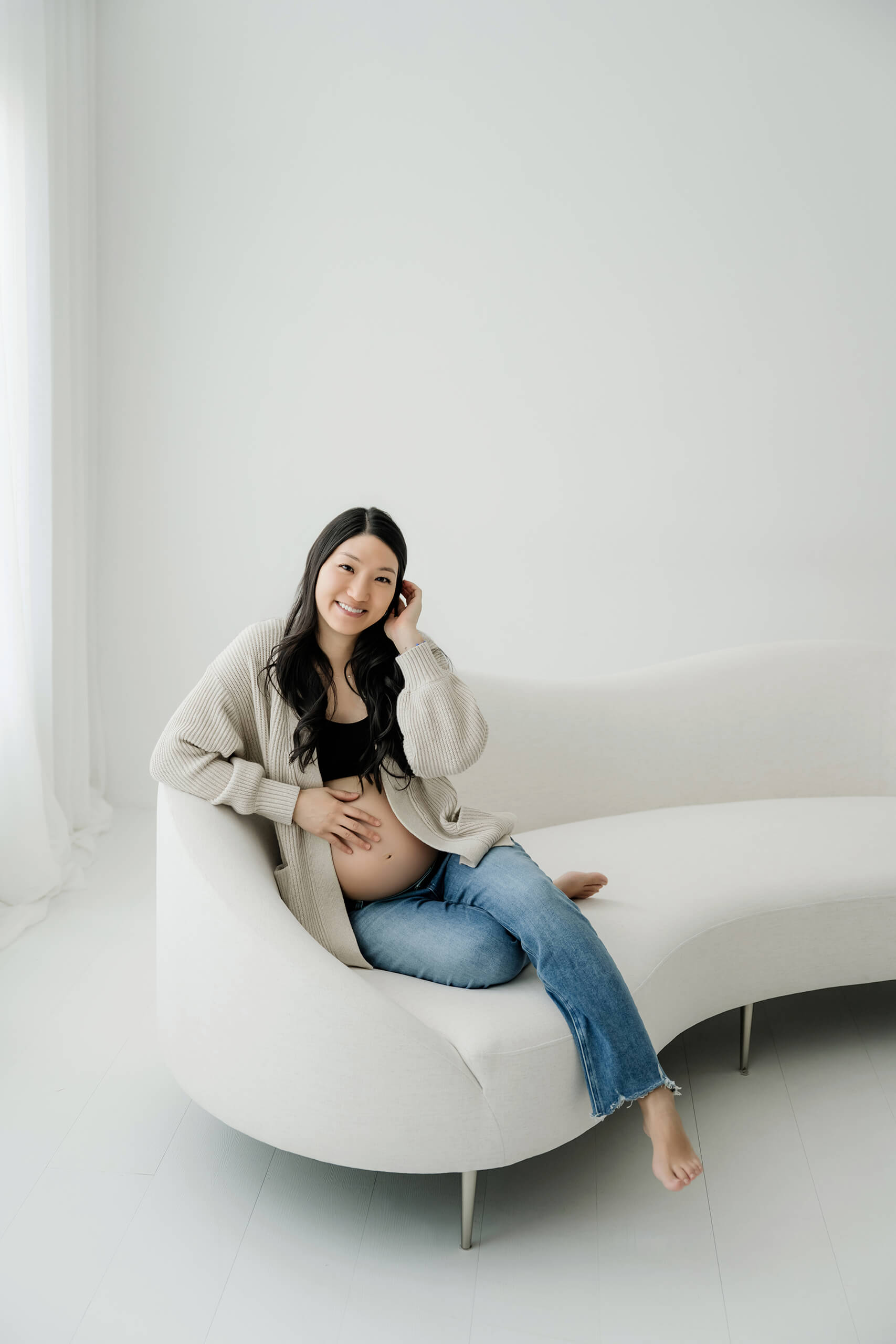 Pregnant woman sitting on a white studio couch with one hand on her belly.
