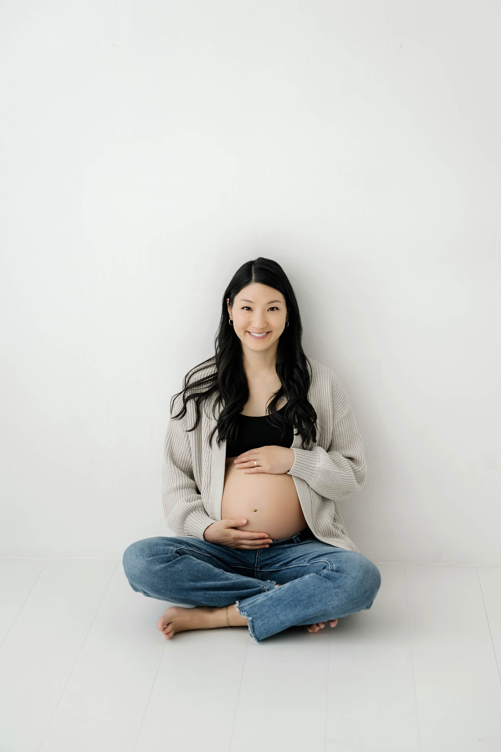 Pregnant woman sitting against a simple white studio wall with her hands on her belly.