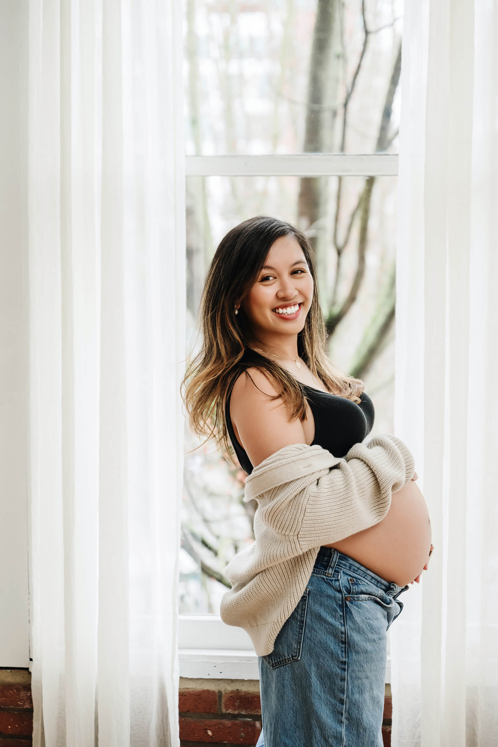 Pregnant woman standing by a large studio window wearing a cardigan and jeans.
