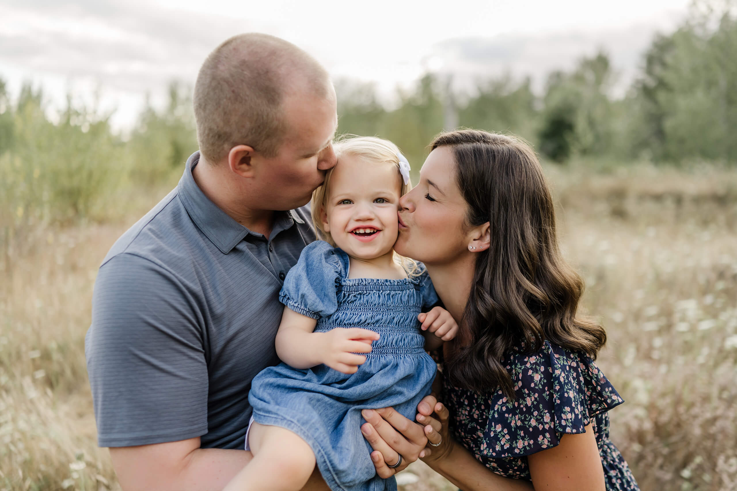 Parents kissing their smiling toddler during an outdoor family photo session in a grassy field.