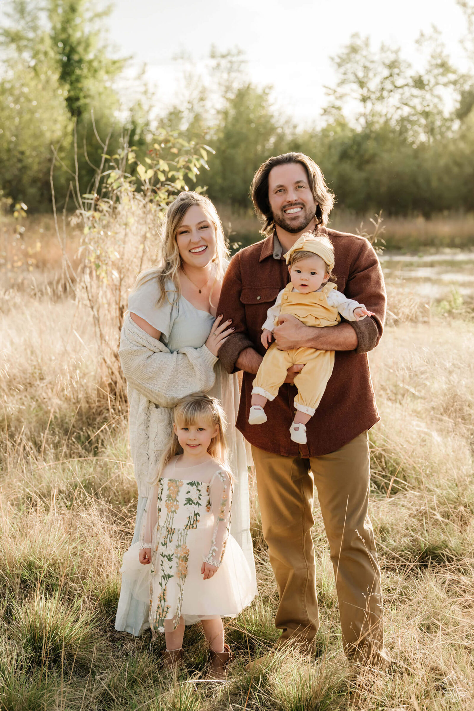 Family of four standing together in a sunlit grassy field during an outdoor family photo session.