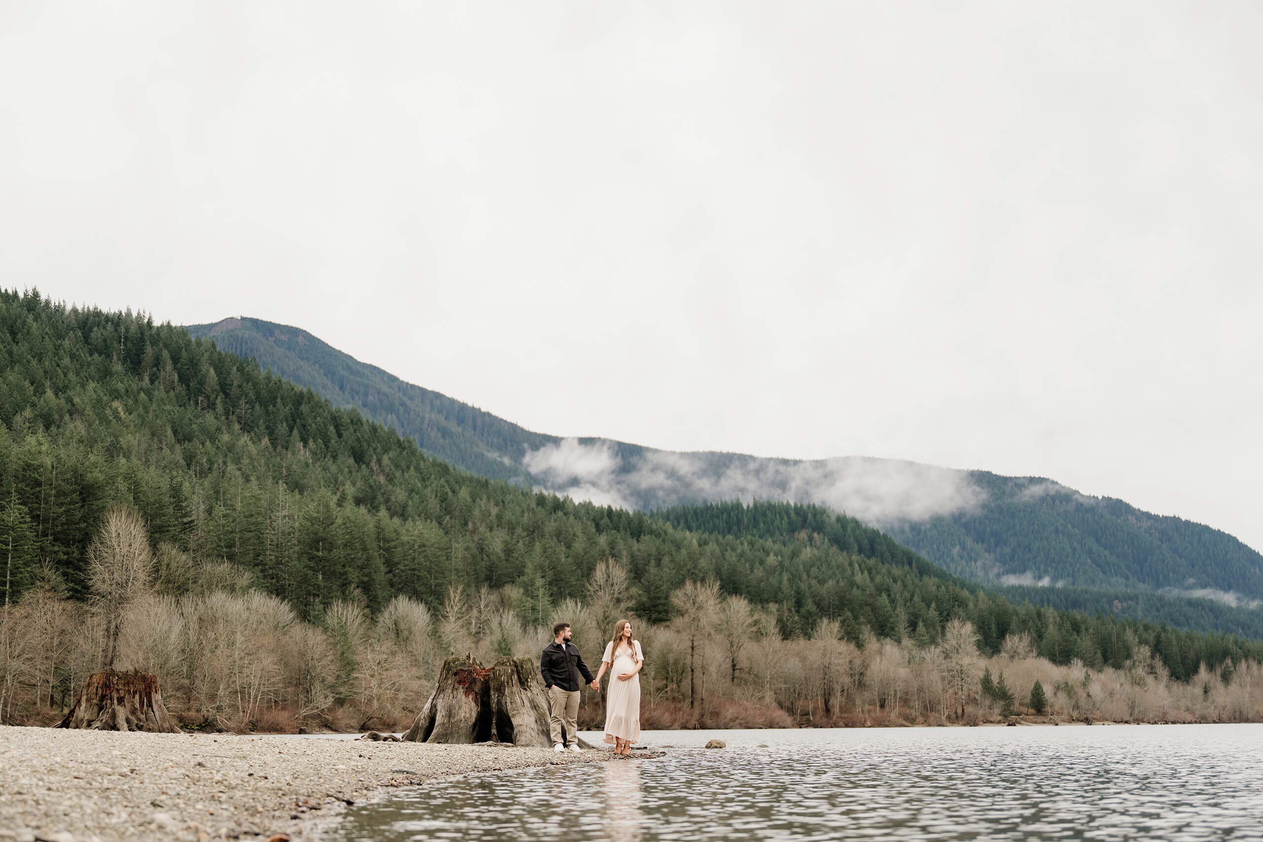 Expecting couple standing by a mountain lake with evergreens and low clouds during an outdoor maternity session.