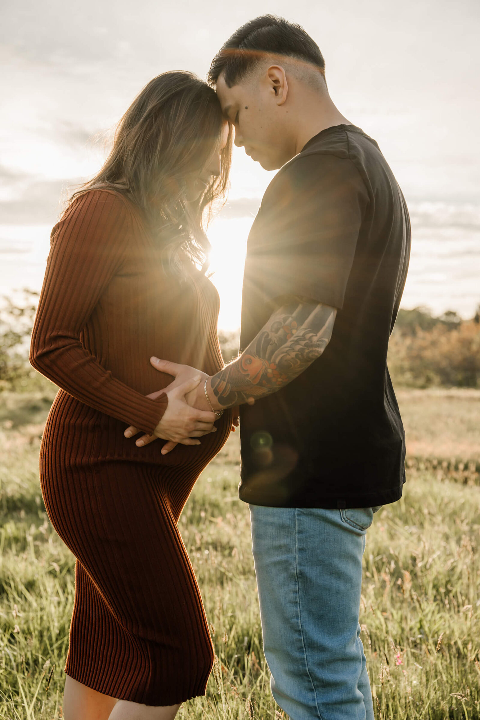Expecting couple holding hands and standing forehead to forehead in a grassy field during a golden-hour maternity session.