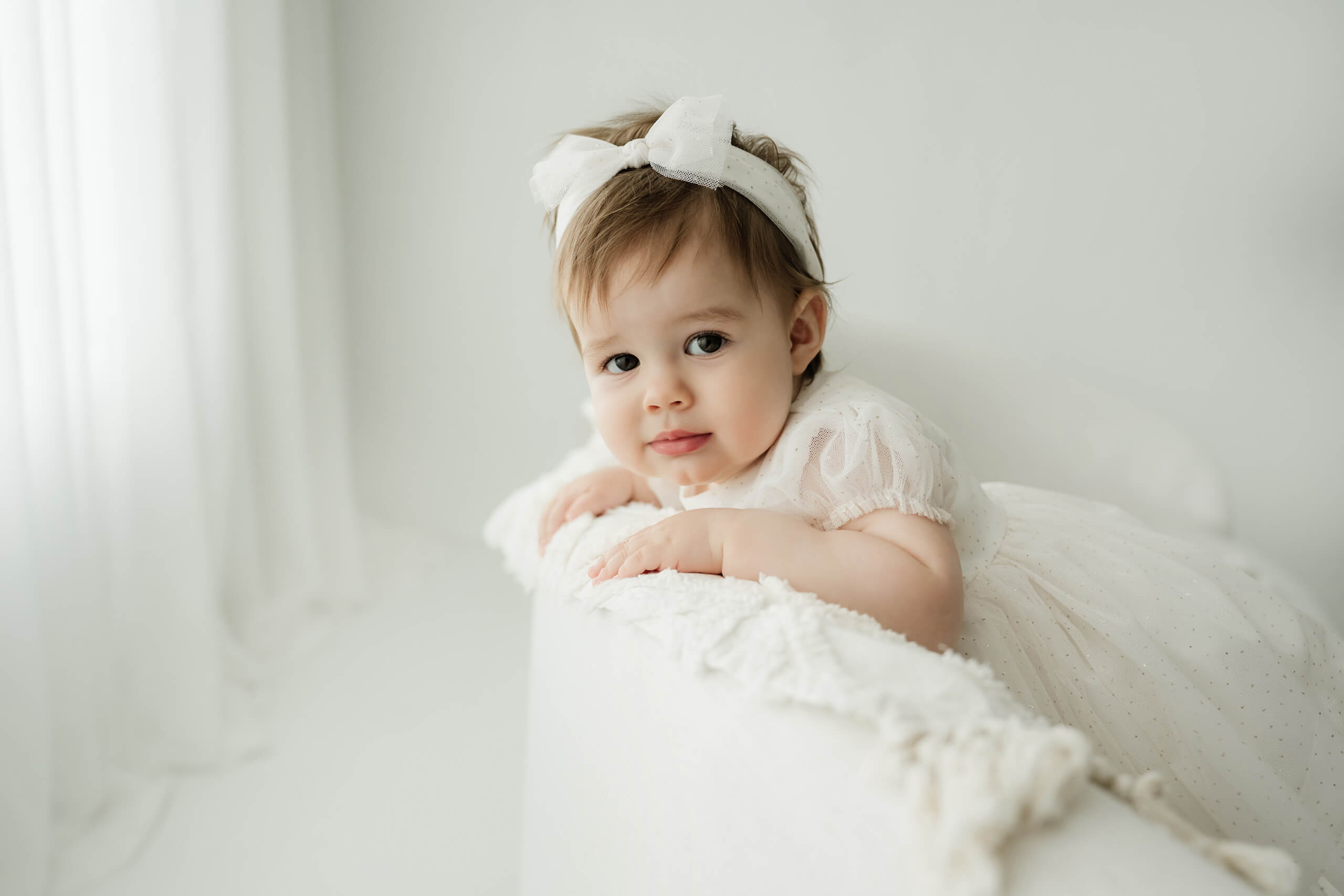 Baby in a white dress and bow posing in a bright studio for a simple milestone portrait.