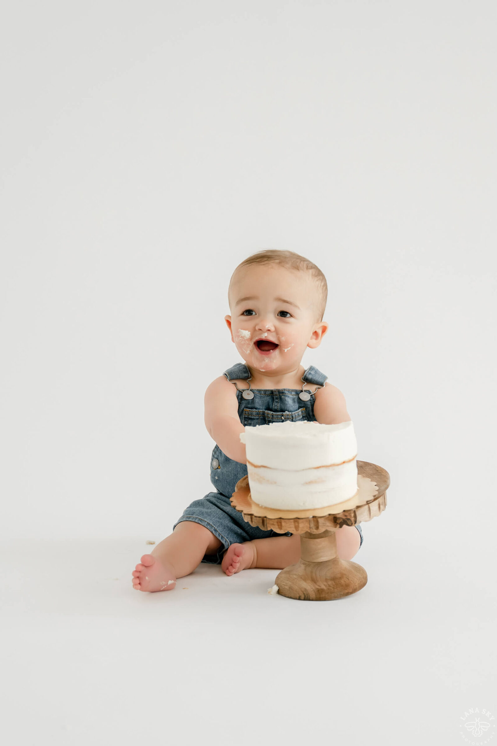 Smiling baby in denim overalls sitting with a simple white birthday cake during a bright studio cake smash session.