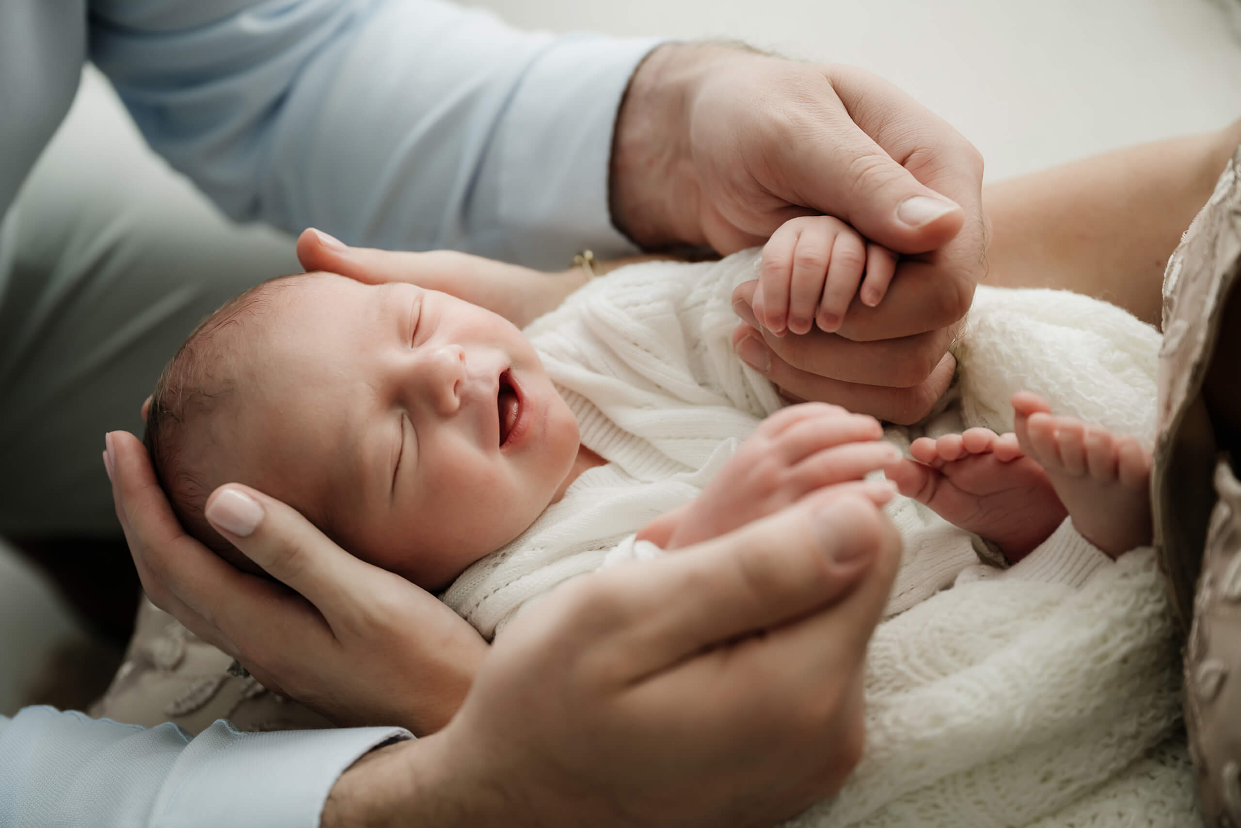 Newborn with eyes closed is cradled in two adults’ hands, with tiny feet and fingers visible.