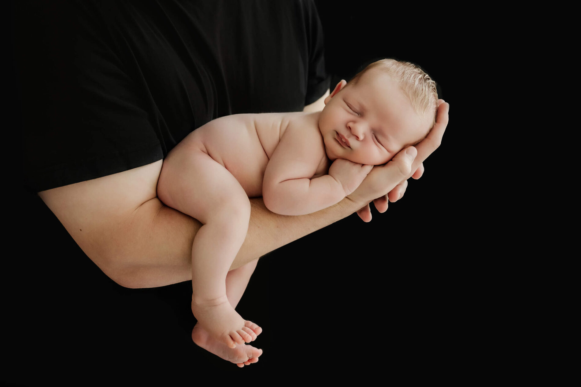 Unwrapped newborn with eyes closed rests across an adult’s forearms against a black background.