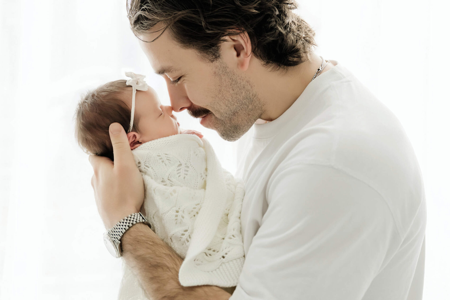 Parent holds a white-wrapped newborn close, touching noses against a bright studio background.