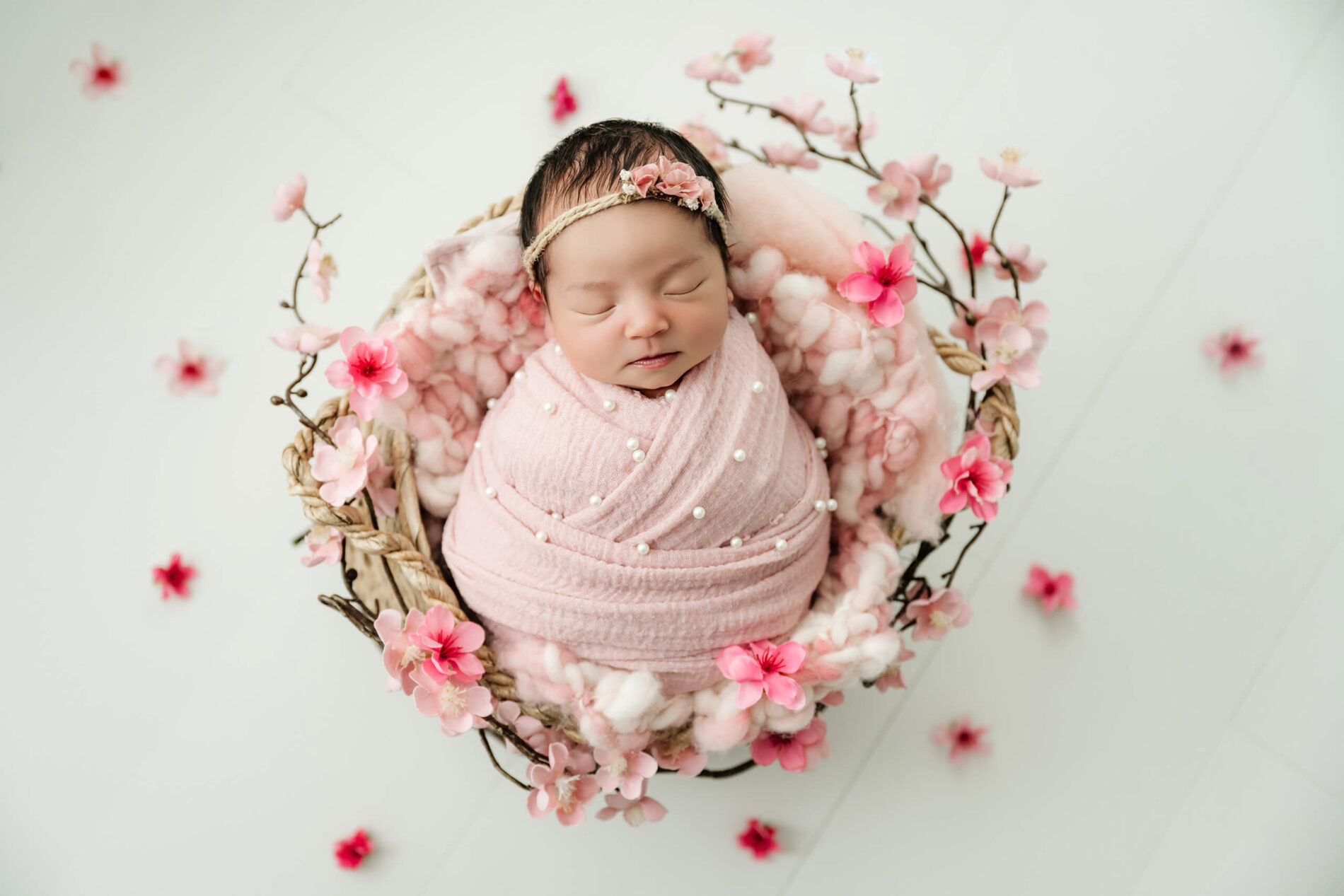 Pink-wrapped newborn with eyes closed lies in a basket surrounded by pink blossoms.
