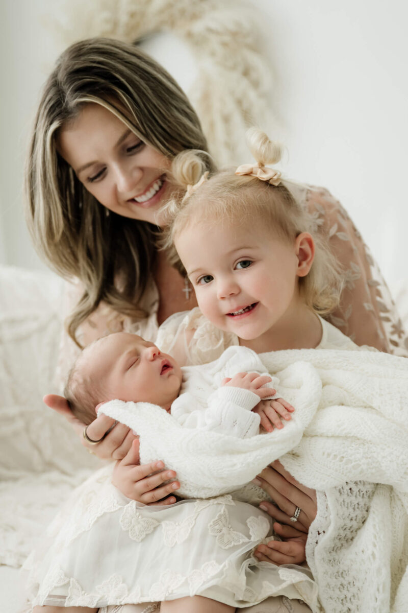 Parent and smiling young sibling cradle a newborn wrapped in a white knit blanket.