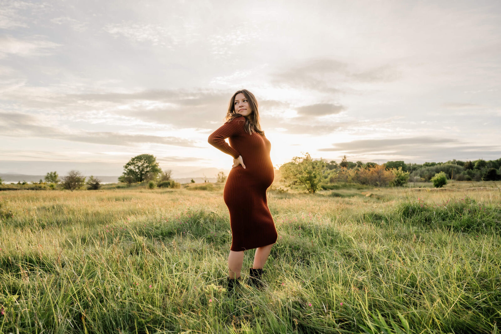 Pregnant woman in a rust-colored dress standing in a grassy Seattle-area field at sunset.