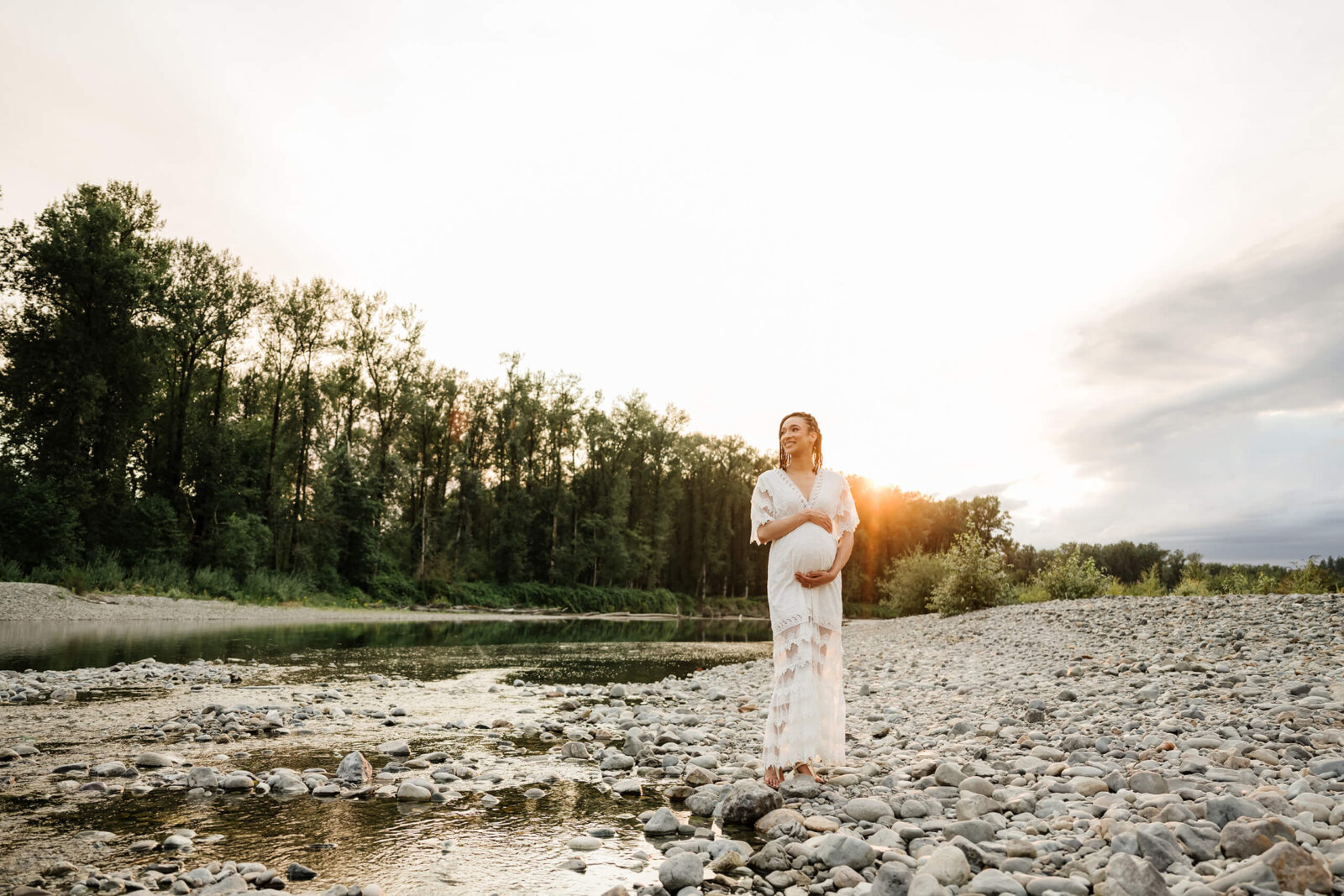 Pregnant woman in a white lace dress standing beside a rocky river in warm evening light.