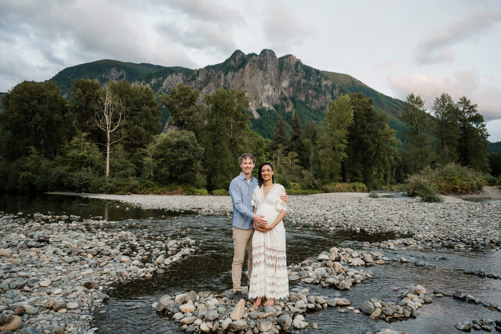 Expecting couple standing on a rocky riverbank with forested mountains behind them.