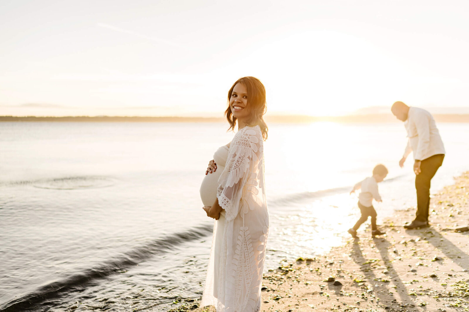Pregnant woman standing by the water while her partner and young child play along the shoreline at sunset.