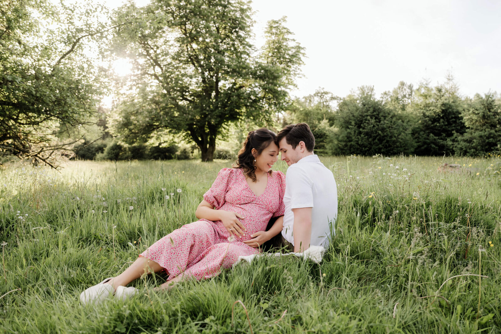Expecting couple sitting together in a grassy field, with the pregnant woman wearing a pink floral dress and white sneakers.