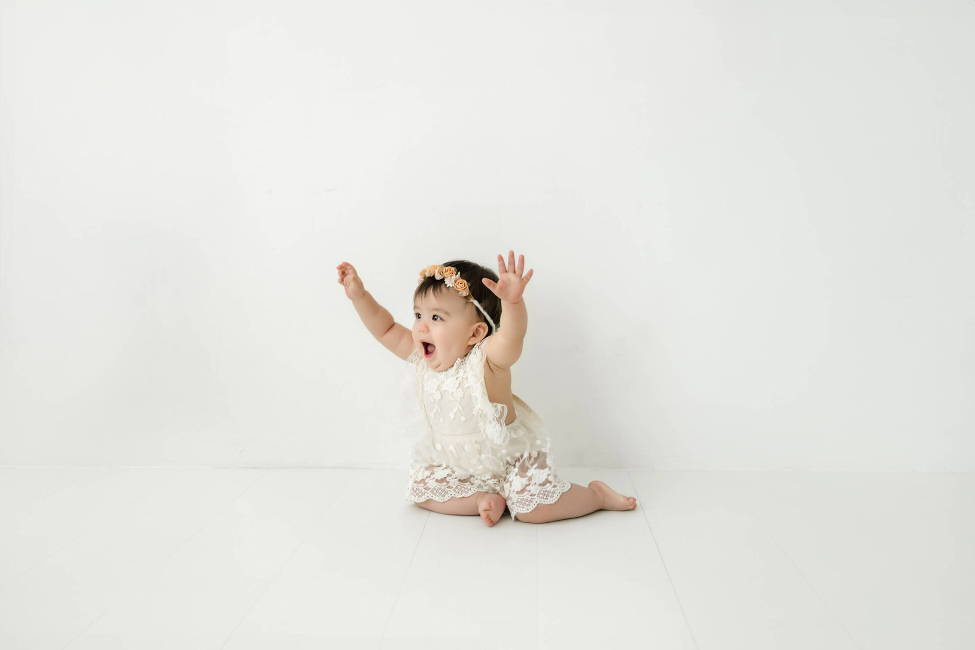 One-year-old baby girl sitting with her arms raised during a Seattle first birthday milestone photography session in a bright white studio.