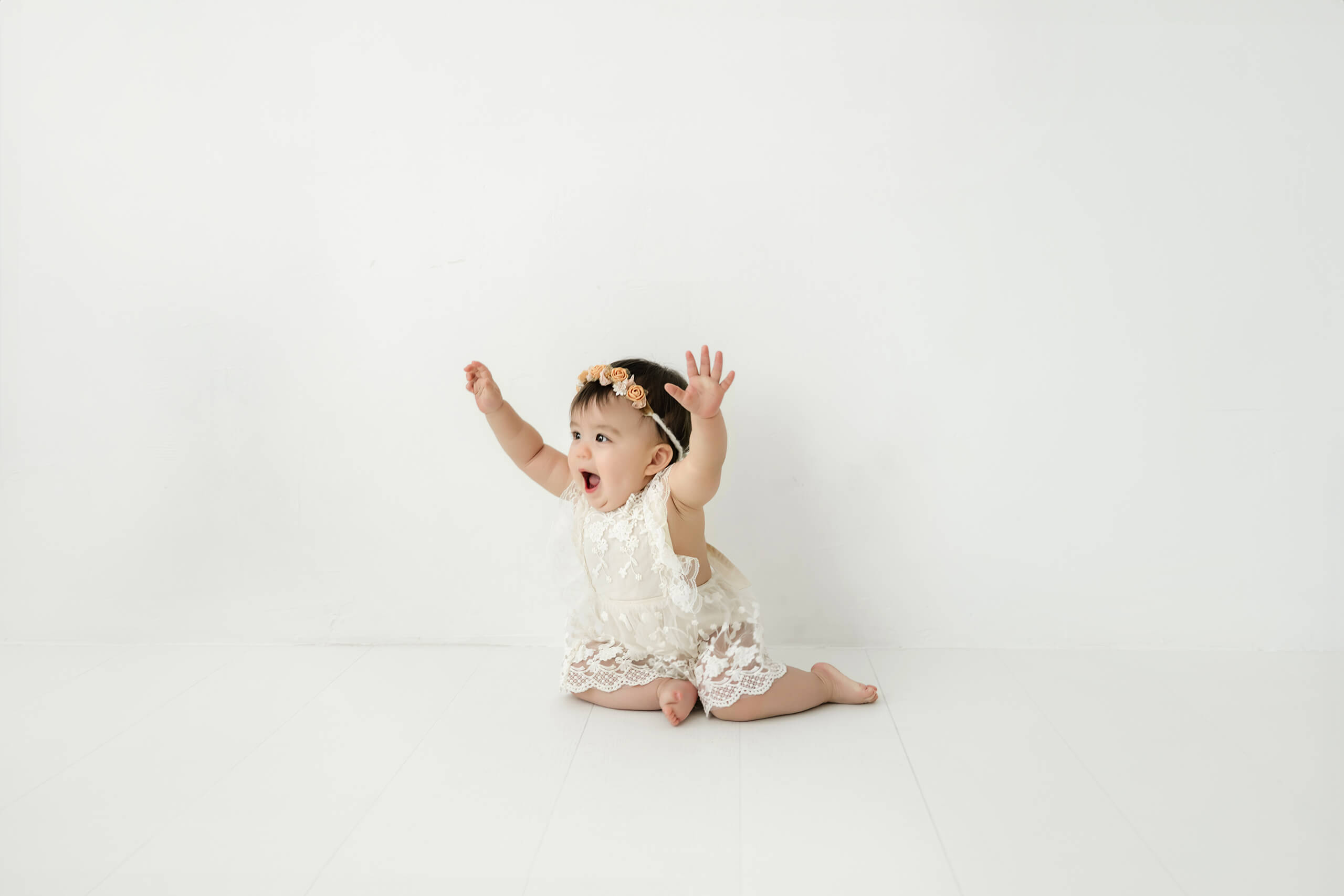 One-year-old baby girl sitting with her arms raised during a Seattle first birthday milestone photography session in a bright white studio.