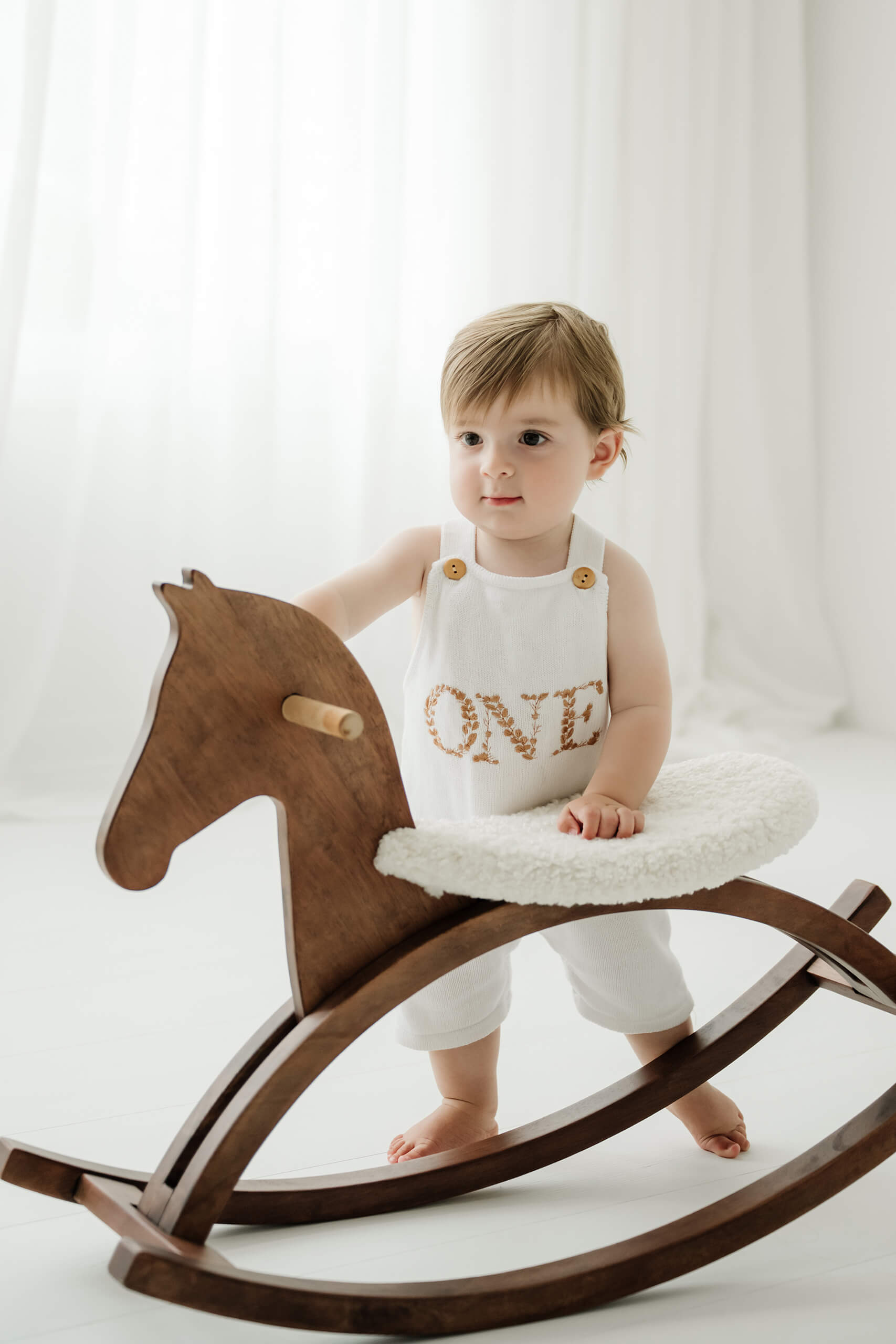 One-year-old boy standing beside a wooden rocking horse during a first birthday milestone portrait session in Kirkland near Seattle.