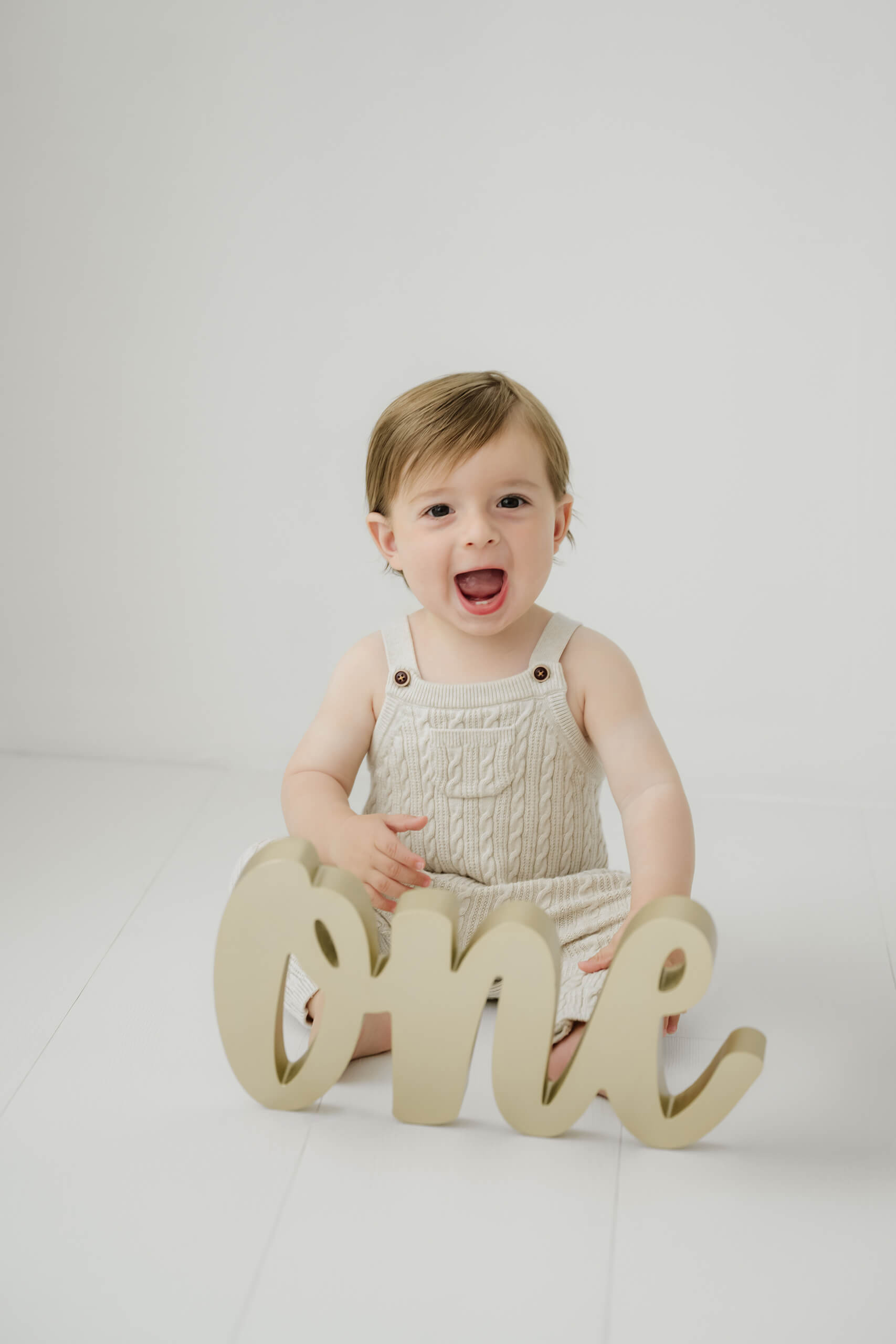 Smiling one-year-old boy with a “one” sign during a Seattle first birthday milestone photography session in a bright white studio.