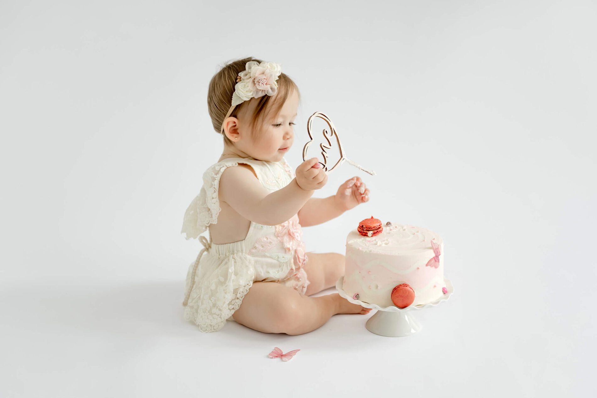 One-year-old girl holding a heart-shaped “one” cake topper beside her pink birthday cake.